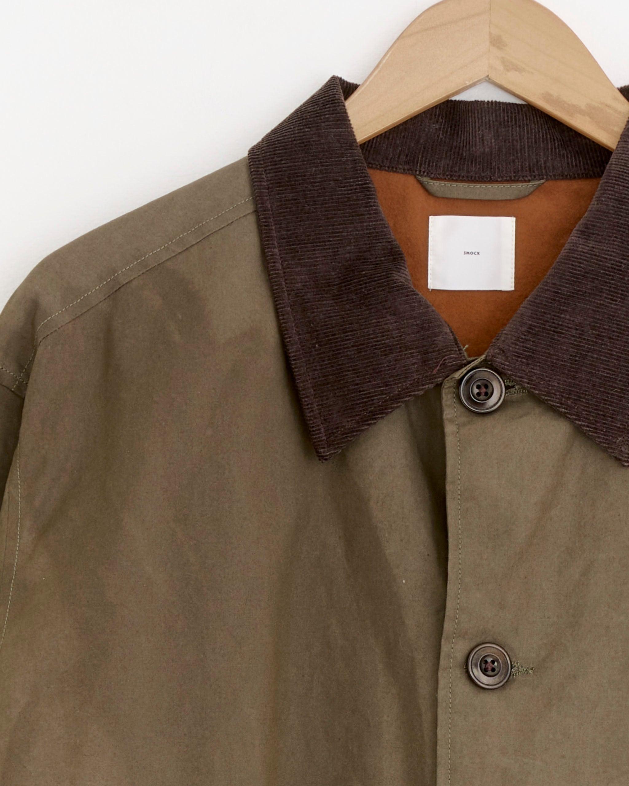 The SMOCK Blakey Jacket in Waxed Cotton Caper features a khaki green finish, brown corduroy collar, large buttons, and a cropped silhouette, shown hanging on a wooden hanger against a white background.
