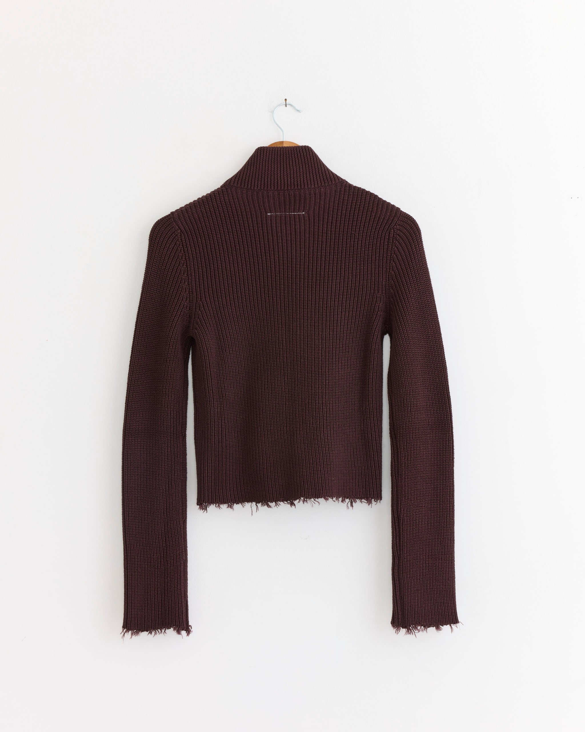 The Zip Cardigan in Ciocolat by MM6 Maison Margiela hangs on a wooden hanger against a white wall, showcasing its long sleeves and raw, unfinished hems.