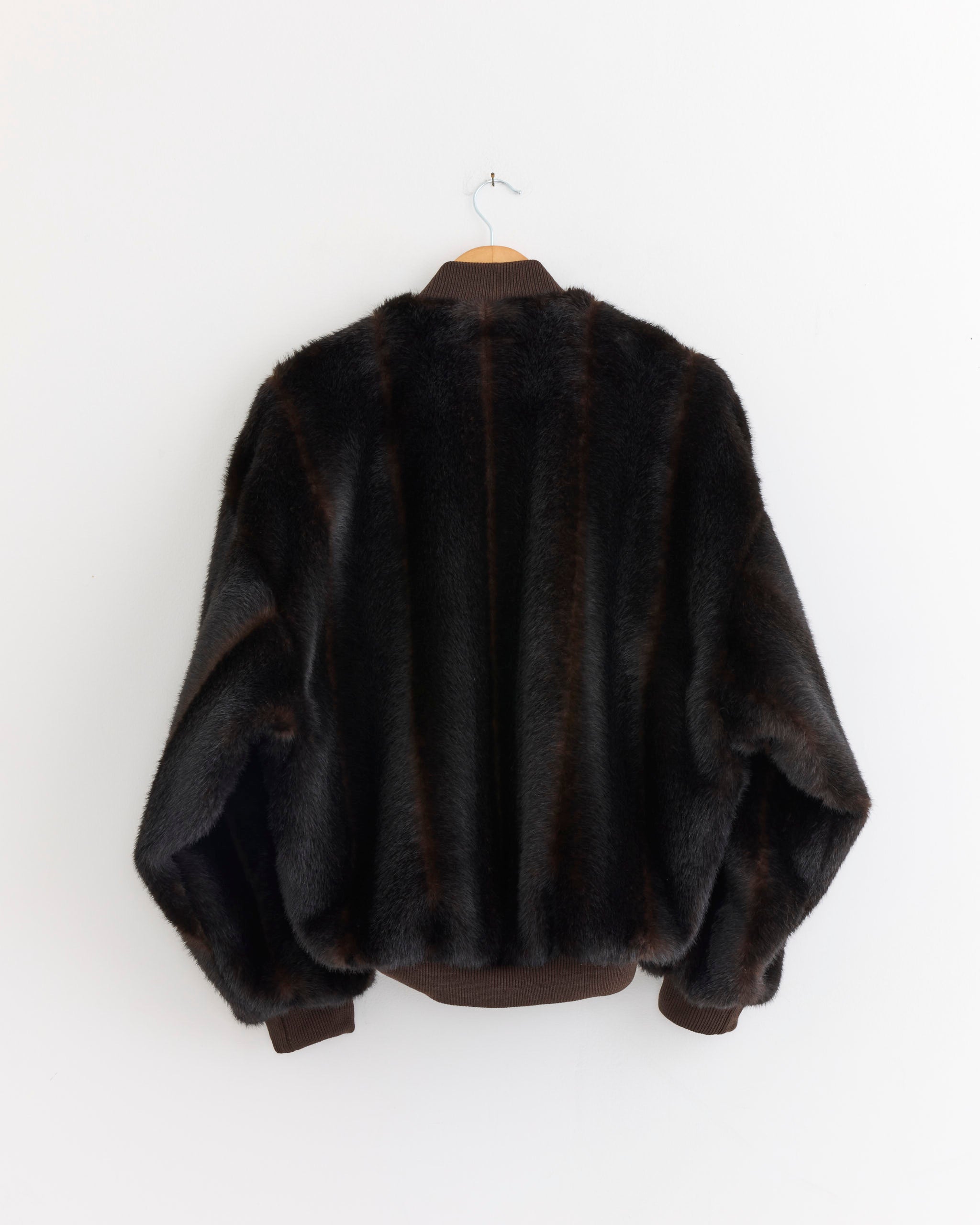 The MM6 Maison Margiela Faux Fur Jacket in Brown, featuring subtle lighter stripes, hangs on a hanger against a plain white wall and is viewed from the back.