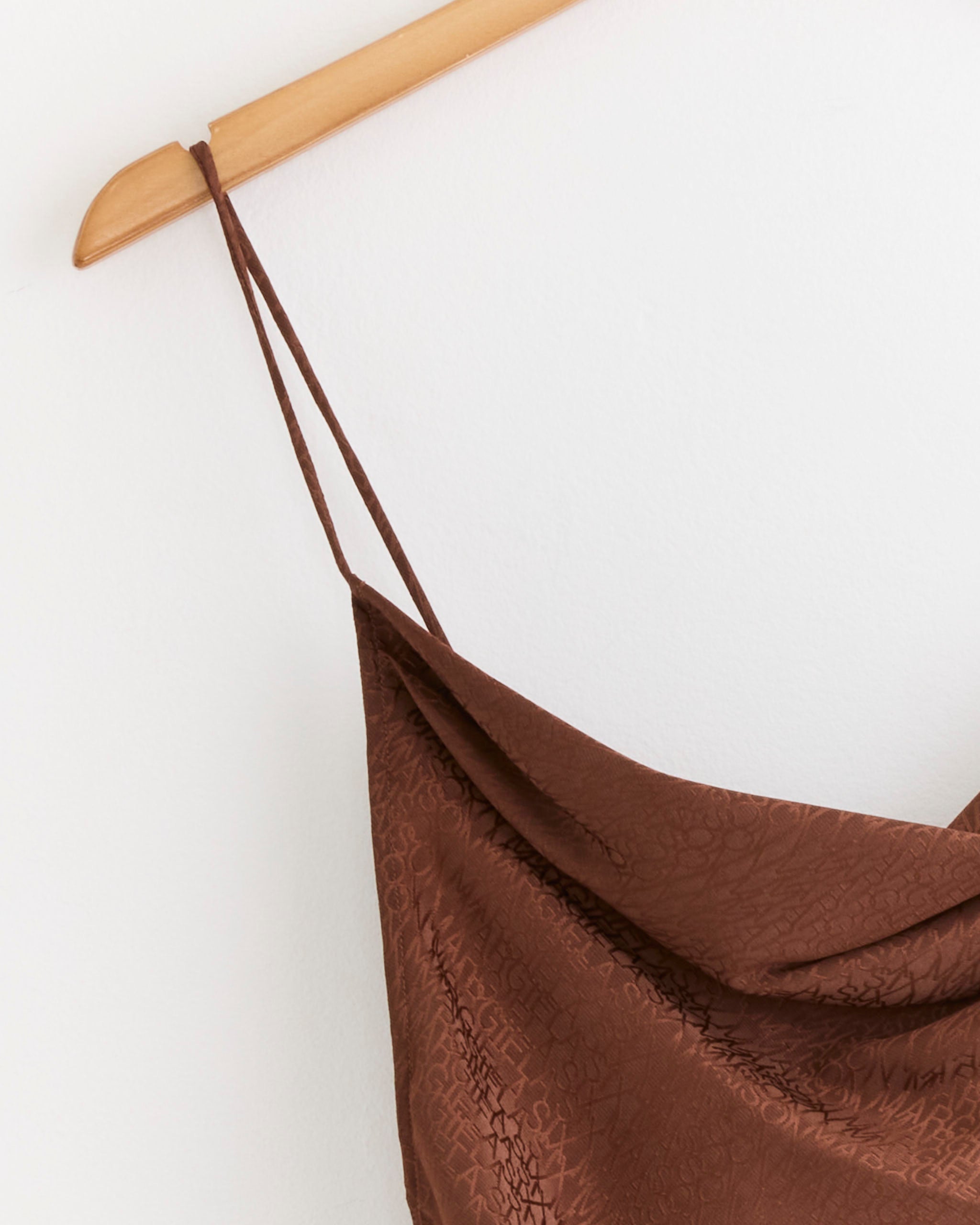 A close-up of the MM6 Maison Margiela Jacquard Top in light brown, featuring textured fabric and thin spaghetti straps, hanging on a wooden hanger against a white wall.