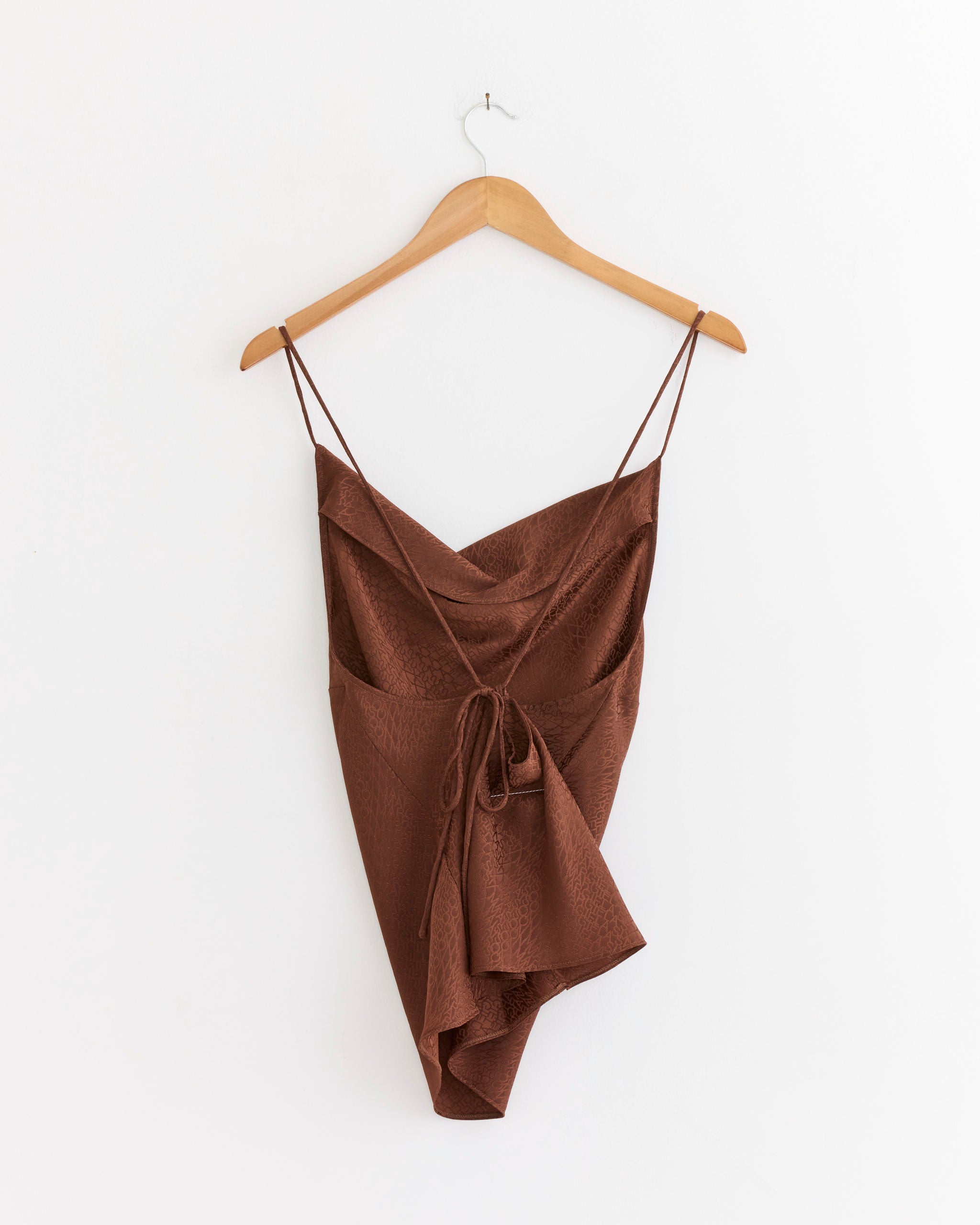 The Jacquard Top in Light Brown by MM6 Maison Margiela, featuring thin spaghetti straps and a front tie detail, hangs on a wooden hanger against a plain white wall.