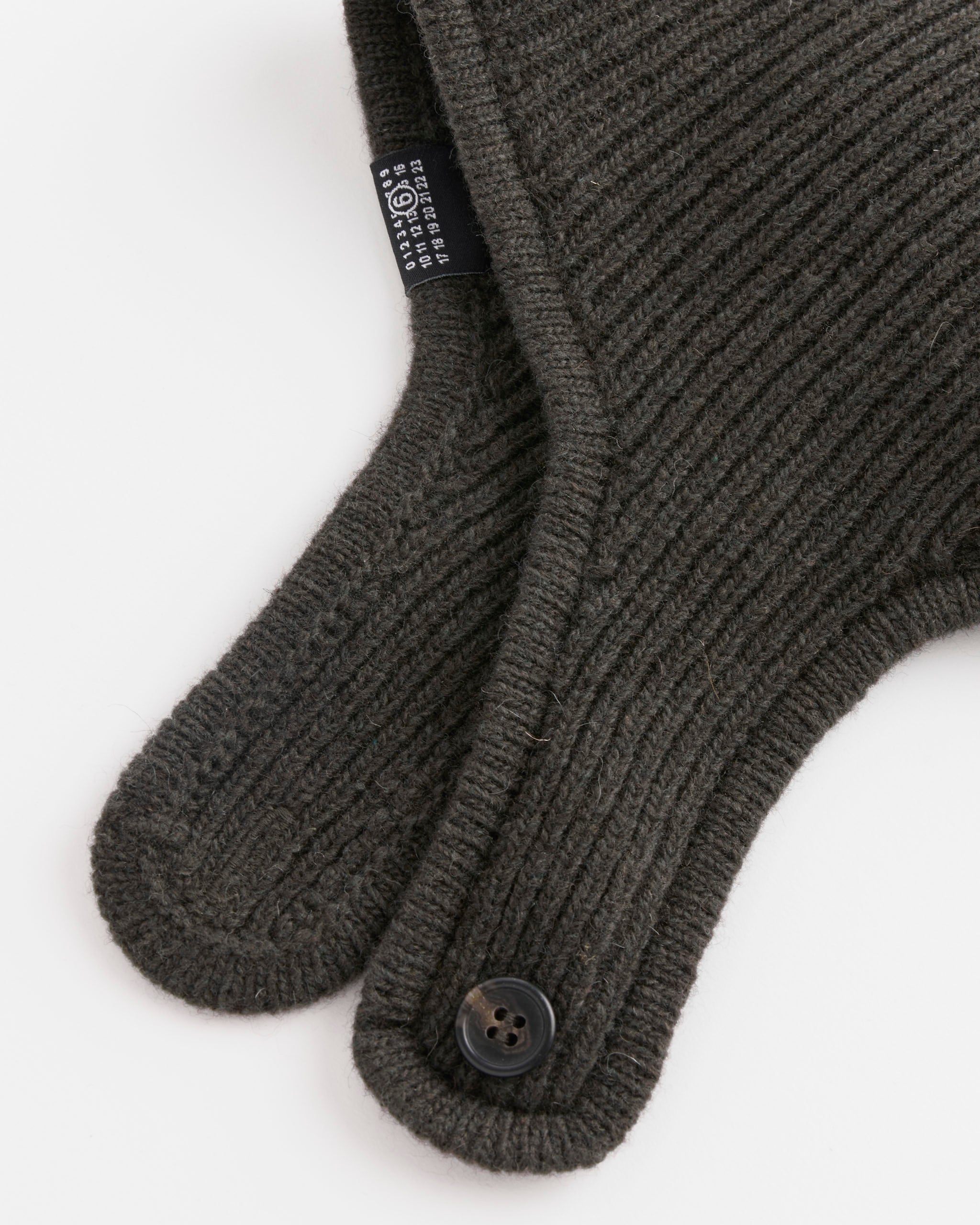 Close-up of MM6 Maison Margiela Beanie in Khaki, featuring overlapping ribbed knit wool straps with buttons. Durable nylon fibers are blended in. A black fabric tag with white text is partially visible.