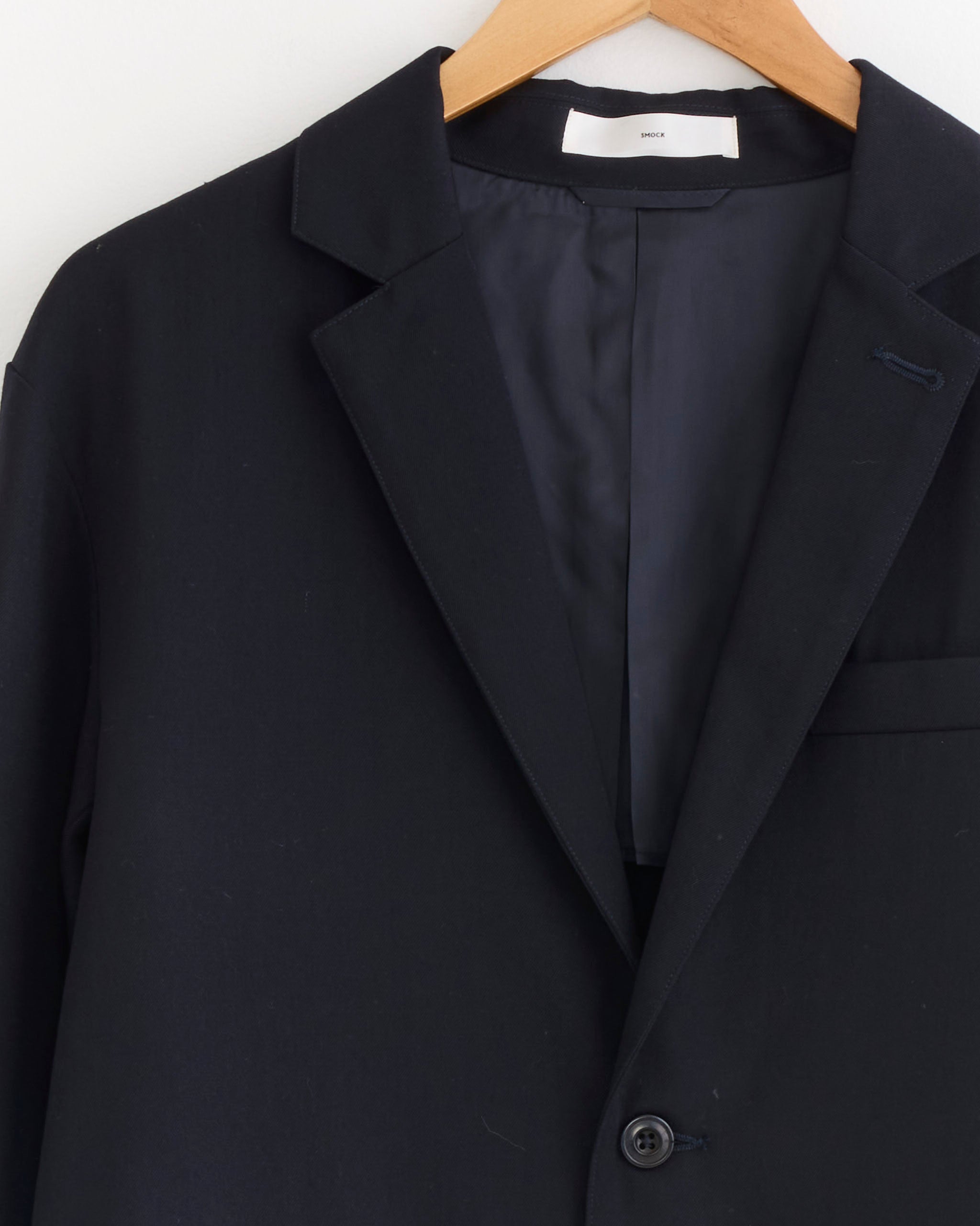 A close-up of the SMOCK Blazer in Wool Gabardine Navy hangs on a wooden hanger, highlighting the lapel, top button, and part of the Bemberg-lined inside pocket against a plain light background.