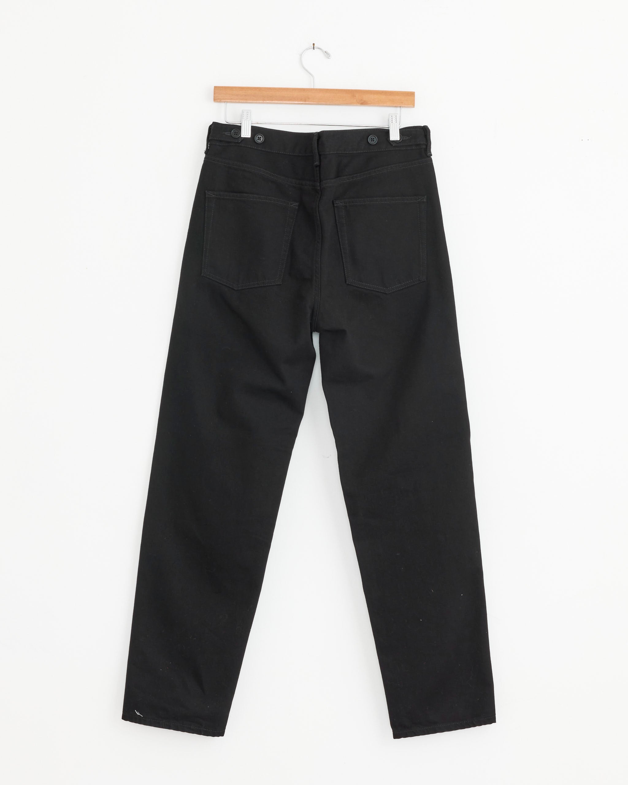 The Tailored Denim Jean in Black by Salon C. Lundman hangs from a wooden hanger against a white background, displayed from the back to show its straight legs, back pockets, and buttoned waistband details.