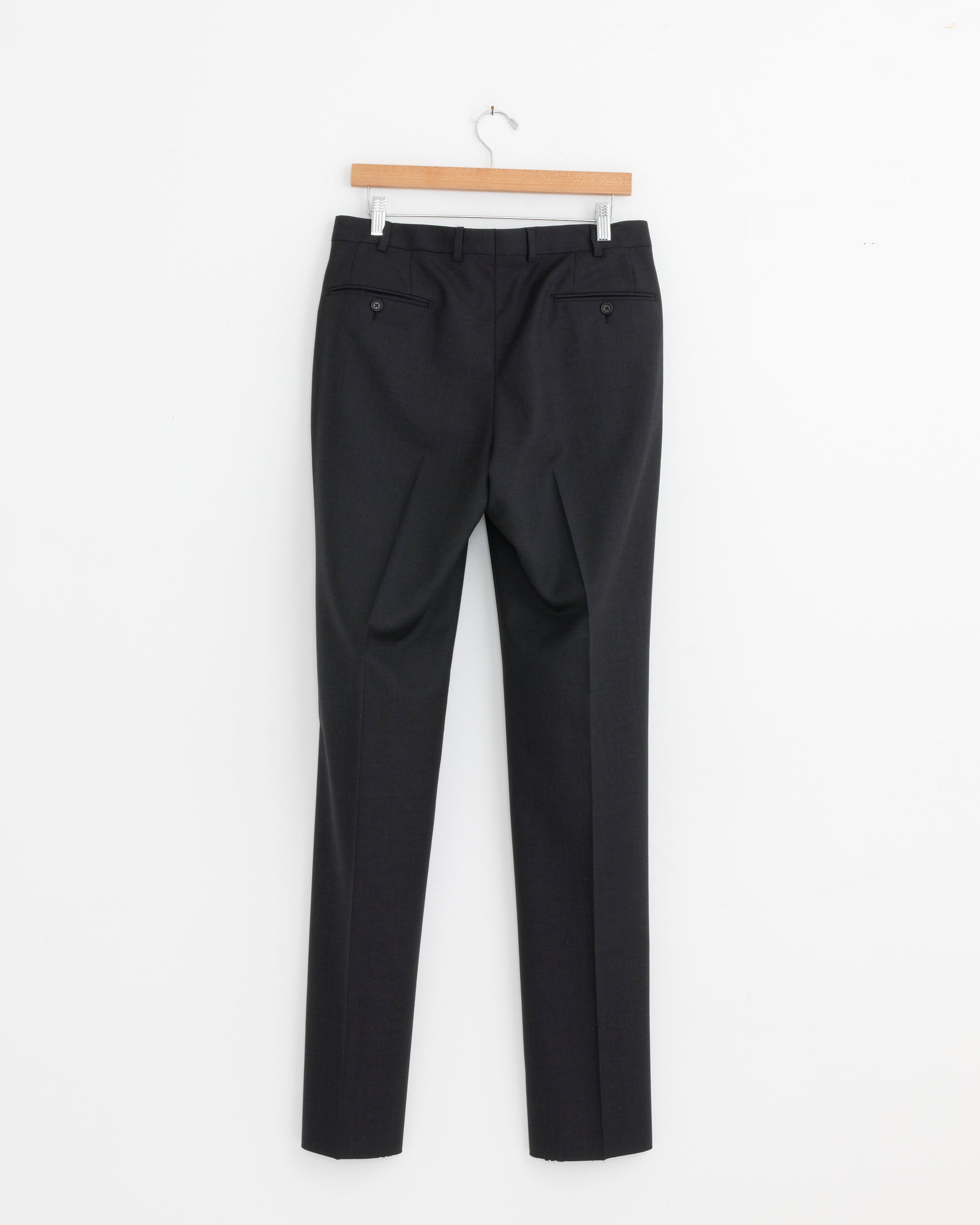 The Salon C. Lundman Double Pleat Trouser in Dark Grey—crafted from 100% wool—hangs on a wooden hanger against a plain white wall, displayed from the back to show its buttoned back pockets.