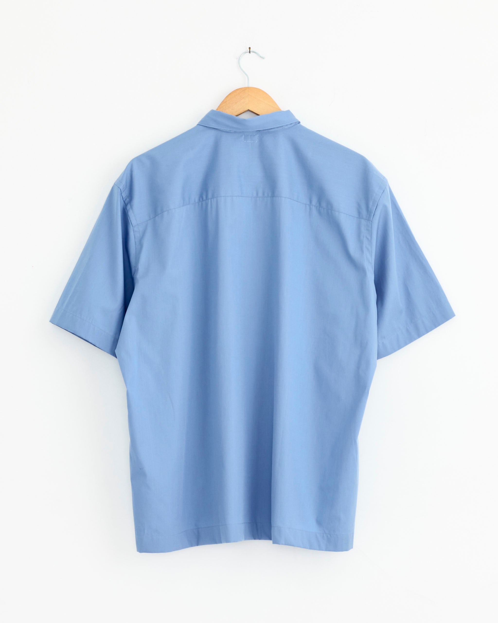 The Necklace Shirt in Clear Blue by Salon C. Lundman, crafted from ItalyAlbini Cotton, features a comfortable fit and refined tailoring. Displayed on a hanger against a white wall, its back view highlights the short sleeves and elegant design.