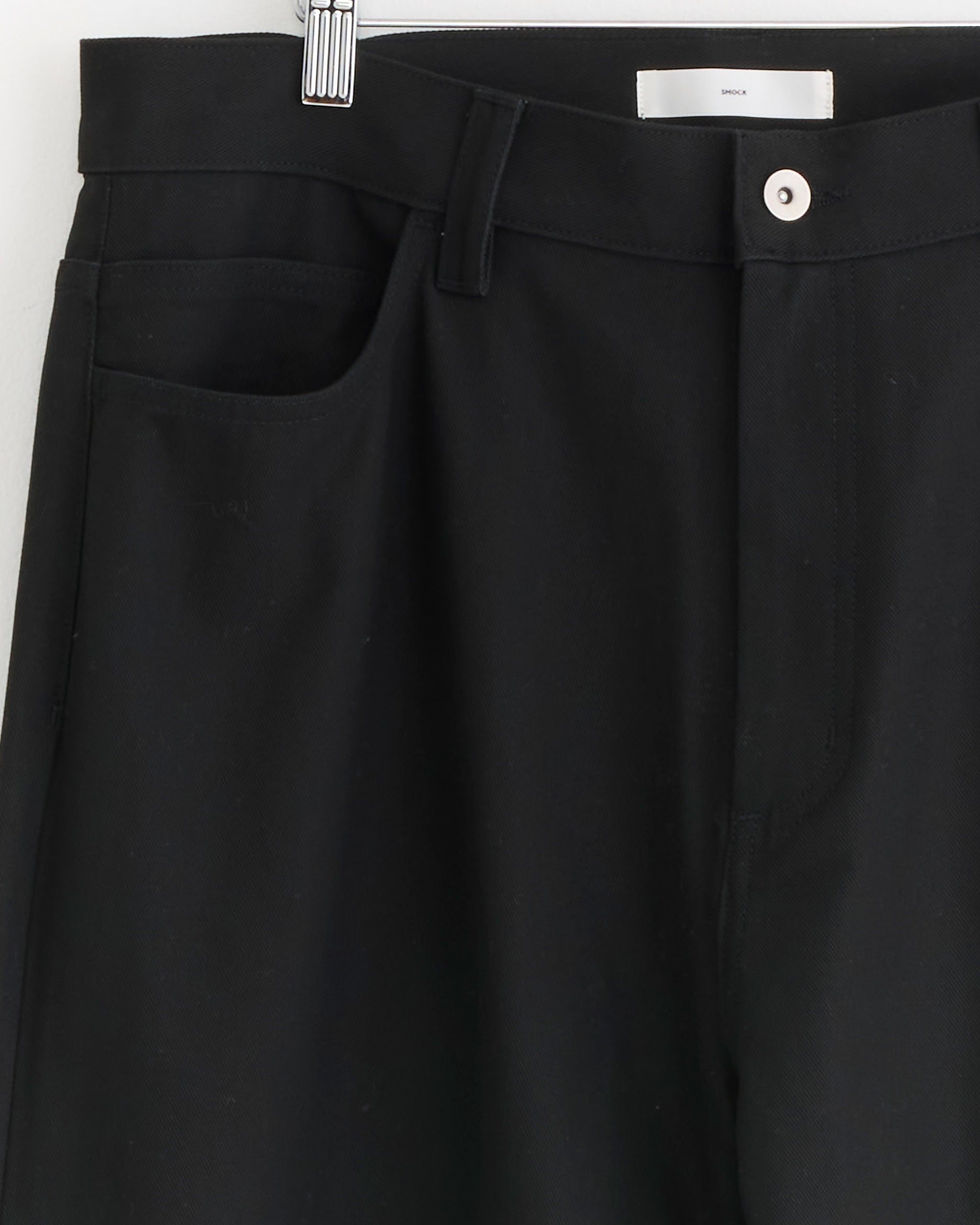 Close-up of the SMOCK Ply Jean in Japanese Cotton Twill Black, featuring the waistband, belt loop, front pocket, and a white button above the zipper while hanging on a hanger.