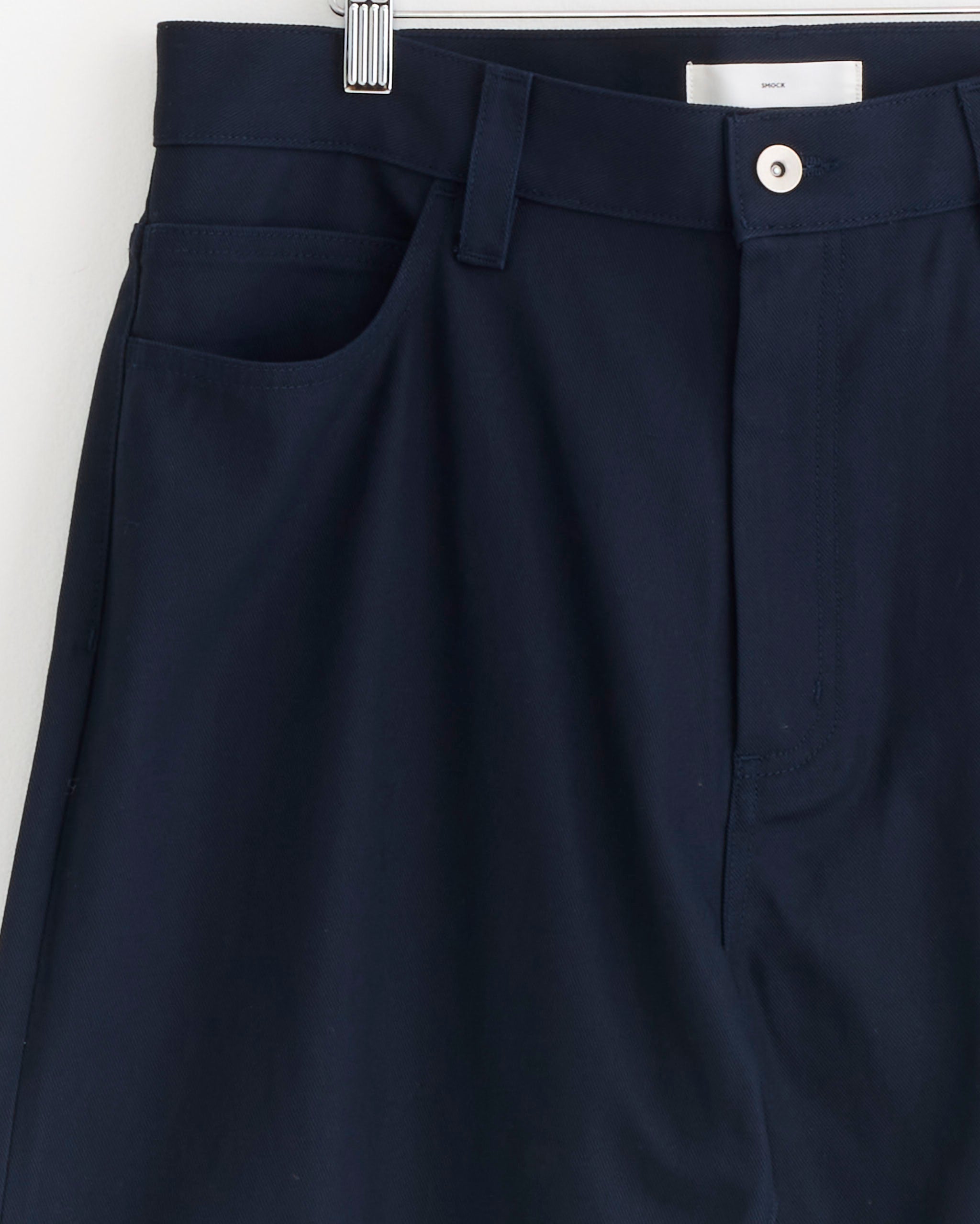 Close-up of the Ply Jean in Japanese Cotton Twill Navy by SMOCK, made in Los Angeles, hanging on a hanger and showing the waistband, front pocket, belt loop, button, and zipper area.