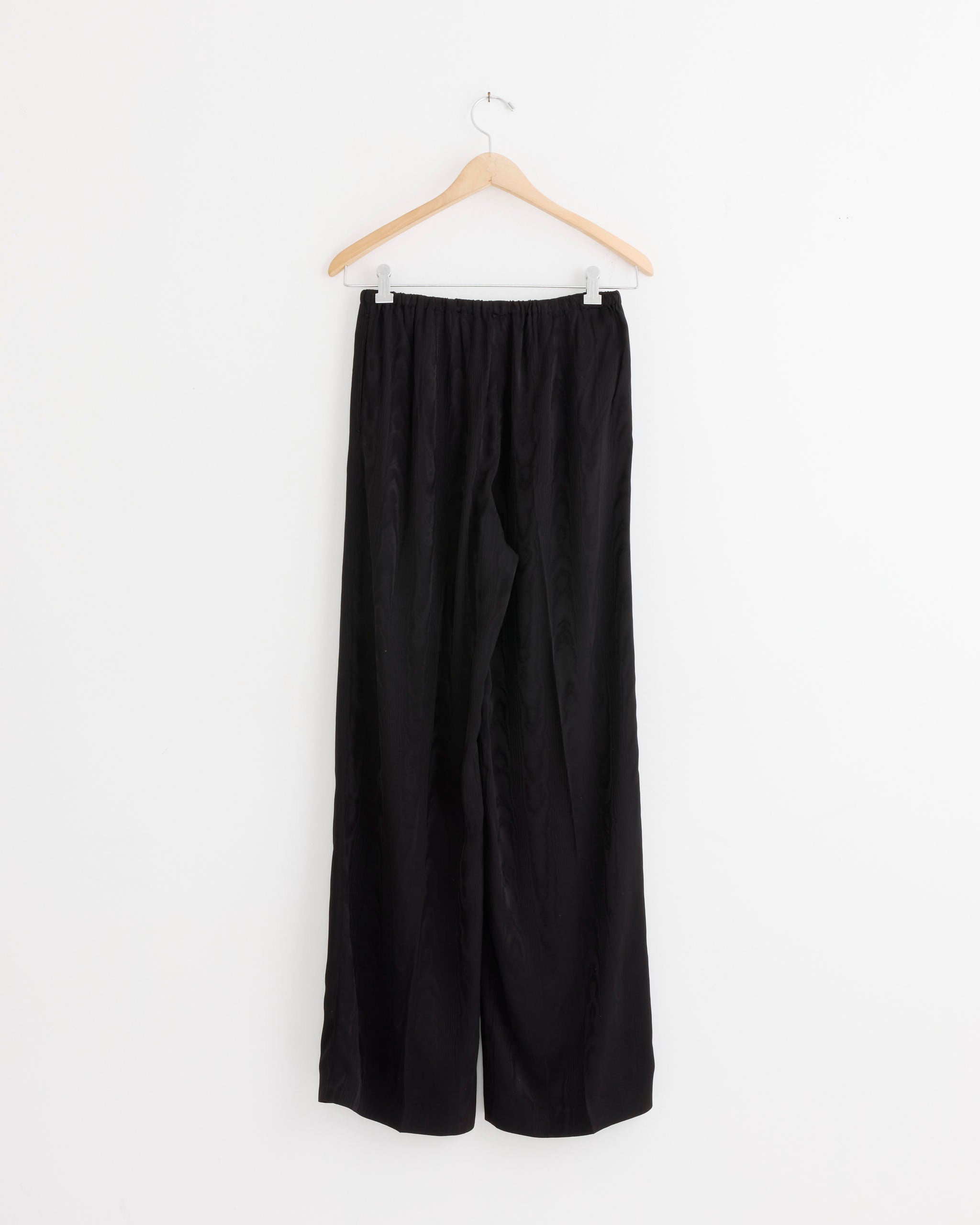 The Dries Van Noten Pant in Black, featuring a relaxed fit and an elastic waistband, crafted from luxurious fabric, is displayed on a wooden hanger against a plain white background.