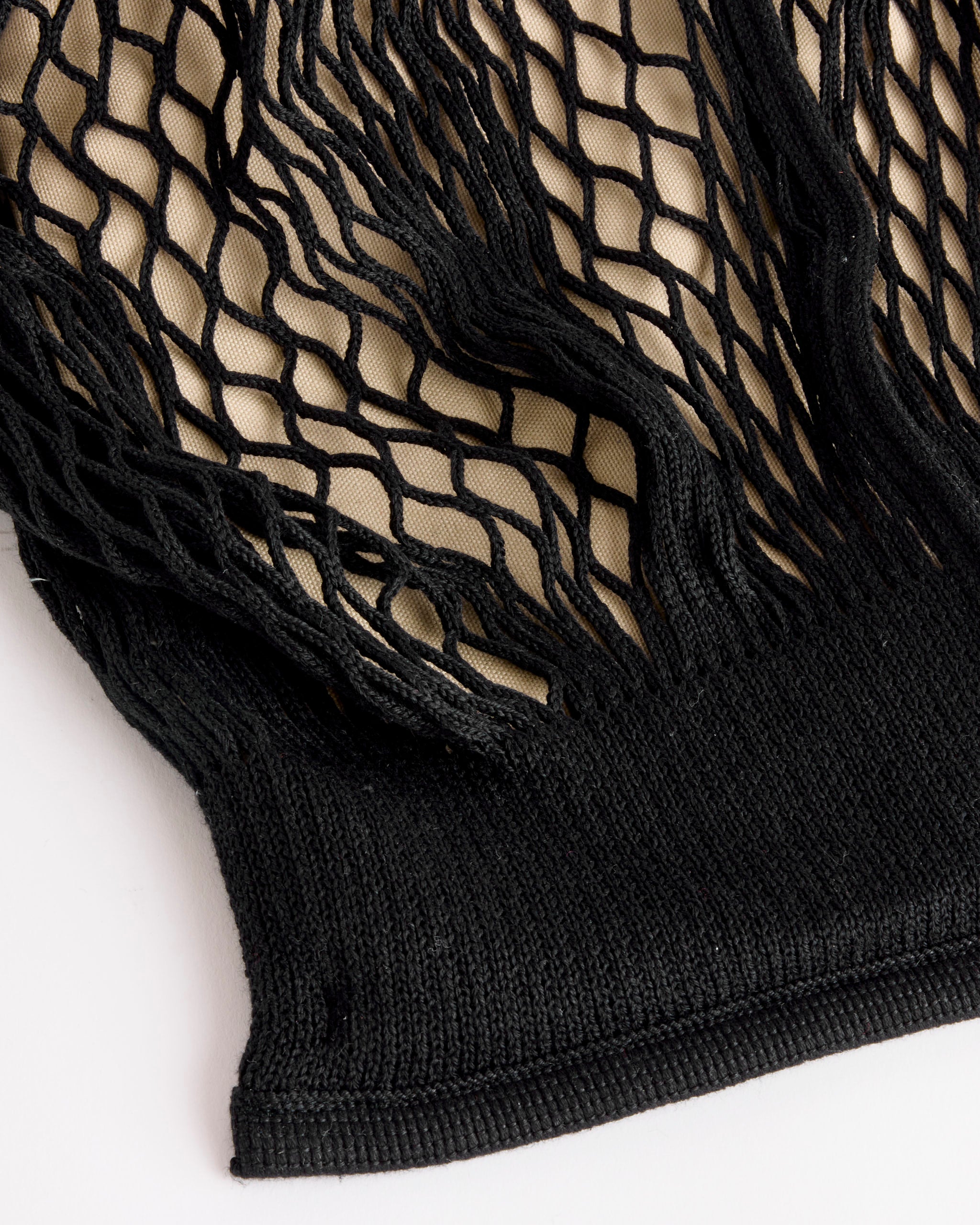 A close-up of Hereu's Calella Filt Black in Black/Natural shows black fishnet fabric with a ribbed edge on a white surface, revealing the beige background beneath.
