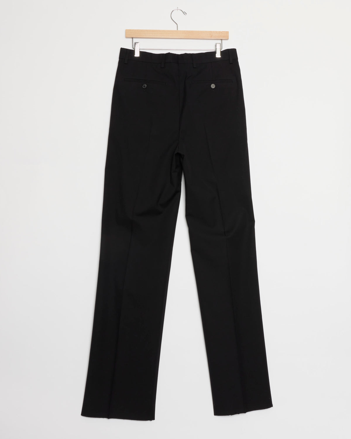 A pair of black pants from the Dries Van Noten Cotton-Wool Suit hangs on a wooden hanger against a plain white background, displayed from the back with two pockets and dark buttons.
