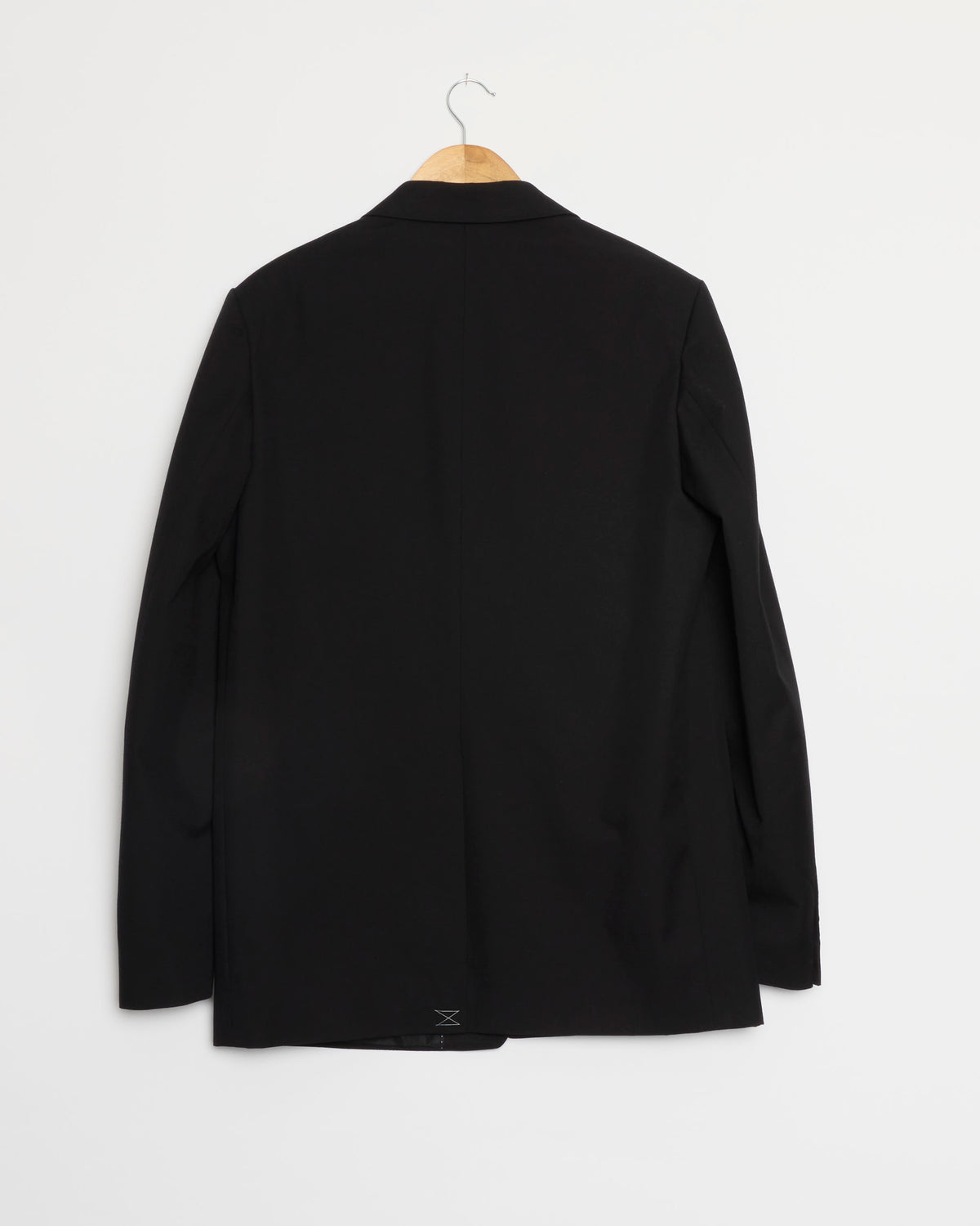 A black Dries Van Noten Cotton-Wool Suit blazer hangs on a wooden hanger against a plain white wall, displayed from the back with long sleeves and a simple, classic design.