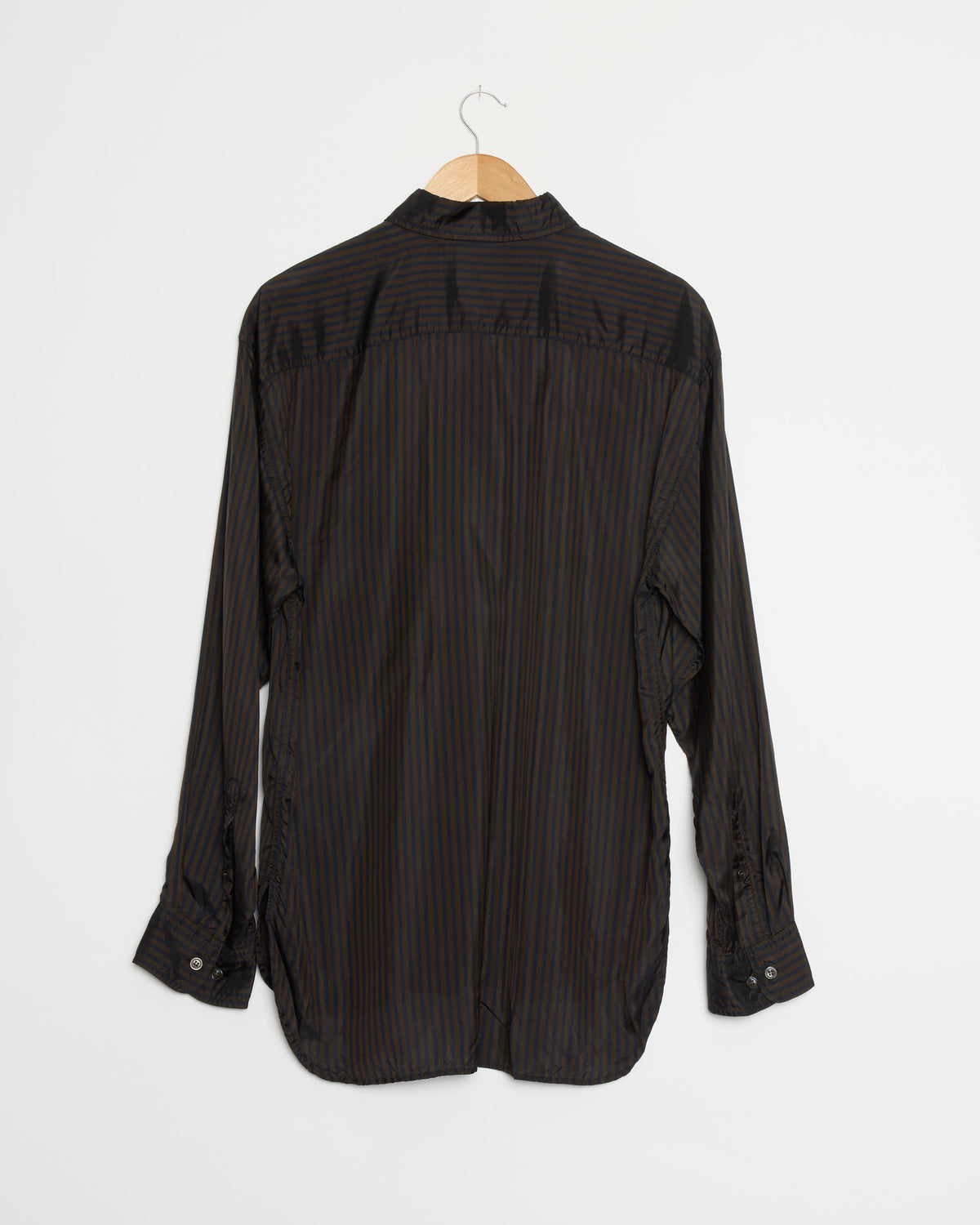 A long-sleeved, striped shirt in dark brown by Dries Van Noten, crafted from luxurious cupro sateen fabric with subtle vertical stripes, hangs on a wooden hanger against a plain white wall and is shown from the back—a style that reflects Dries Van Noten’s refined aesthetic.