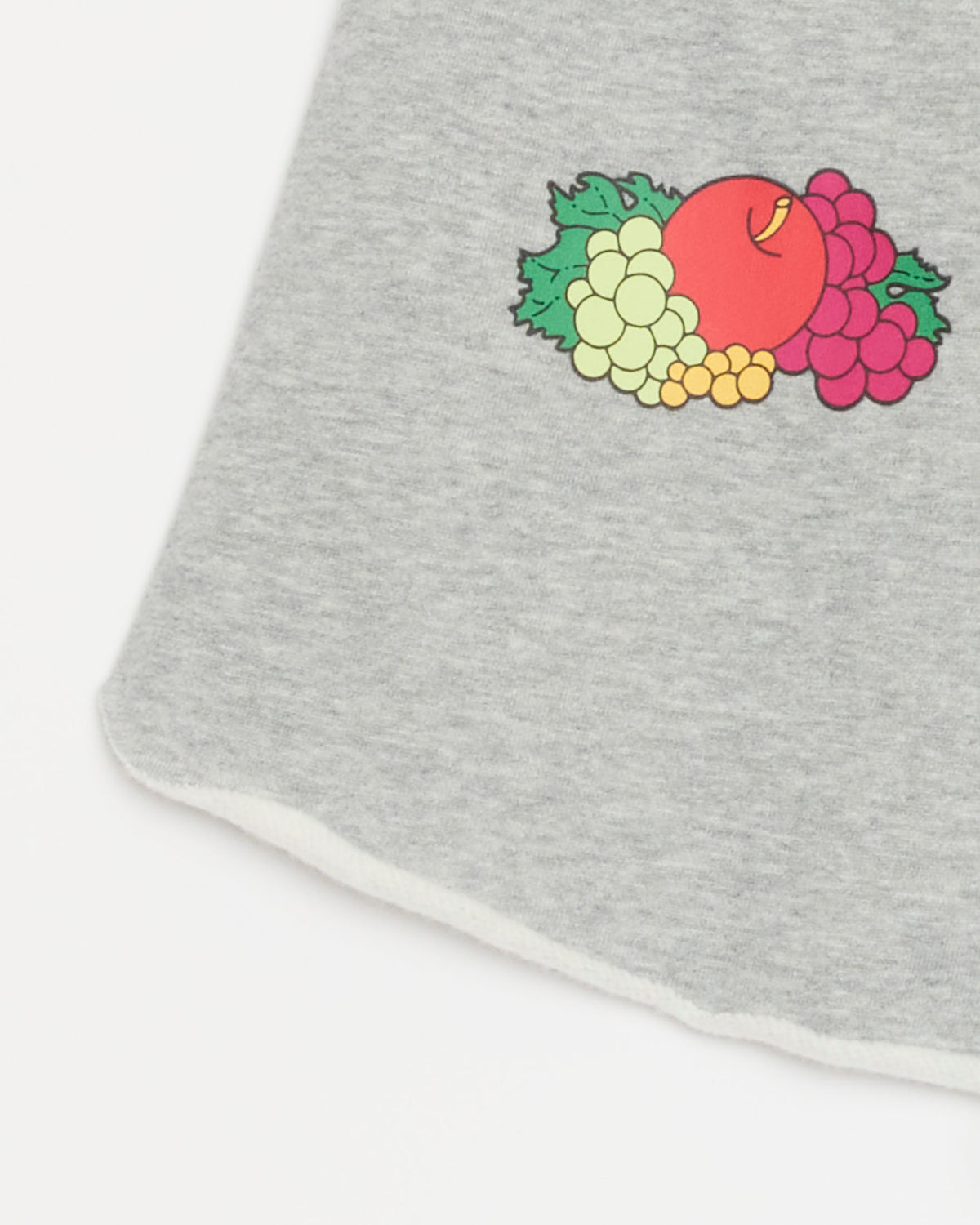 A close-up of the Hommegirls Fruity Short in Heather Grey showcases its soft gray fabric and vibrant fruit graphic—including a red apple, green and purple grapes, and lush green leaves—printed near the edge. These vintage-inspired shorts resemble cutoff sweatpants, offering playful style and laid-back comfort.