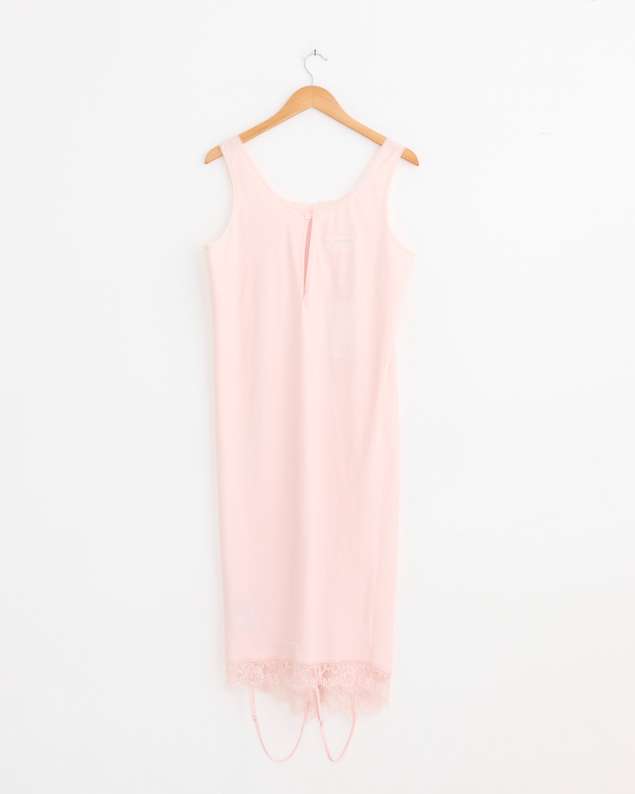 The Mirrored Lace Slip Dress in Pink by MM6 Maison Margiela, featuring a sleeveless design and delicate lace details at the neckline and hem, hangs on a wooden hanger against a plain white wall.