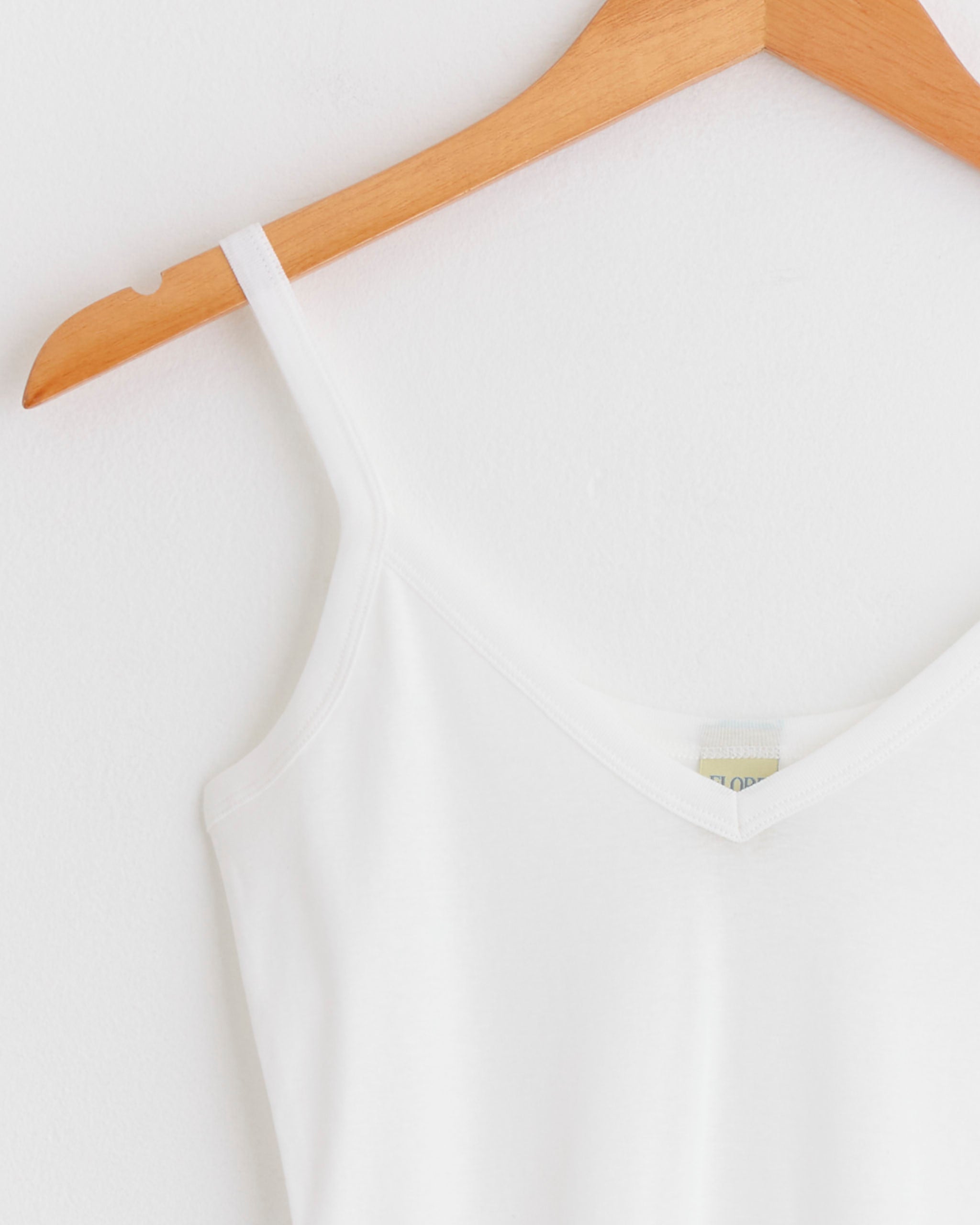 The Isla Cami in White by FLORE FLORE, a spaghetti strap tank top, hangs on a wooden hanger against a plain white wall.