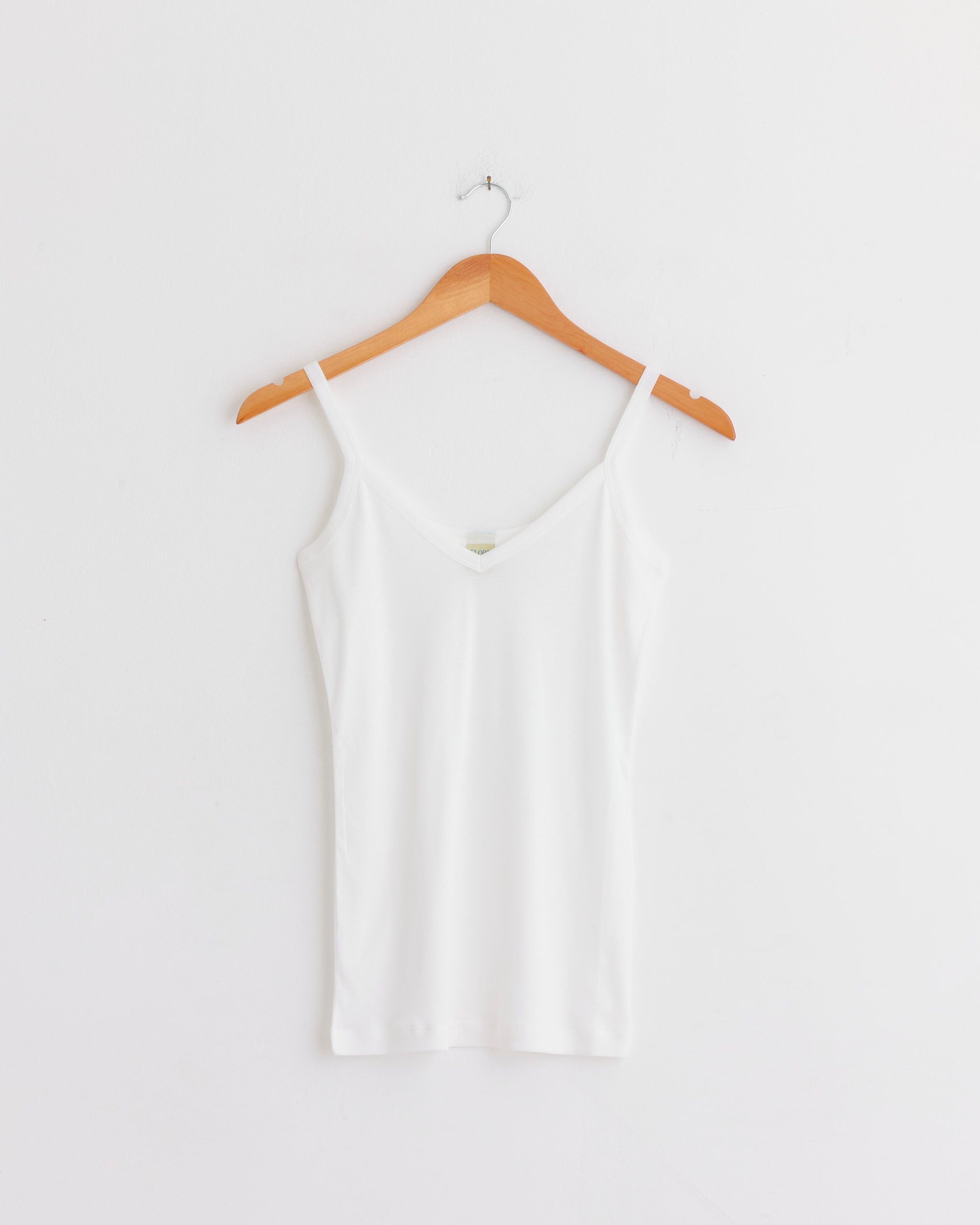 The FLORE FLORE Isla Cami in White, a simple spaghetti strap tank top, is displayed on a wooden hanger against a white wall.
