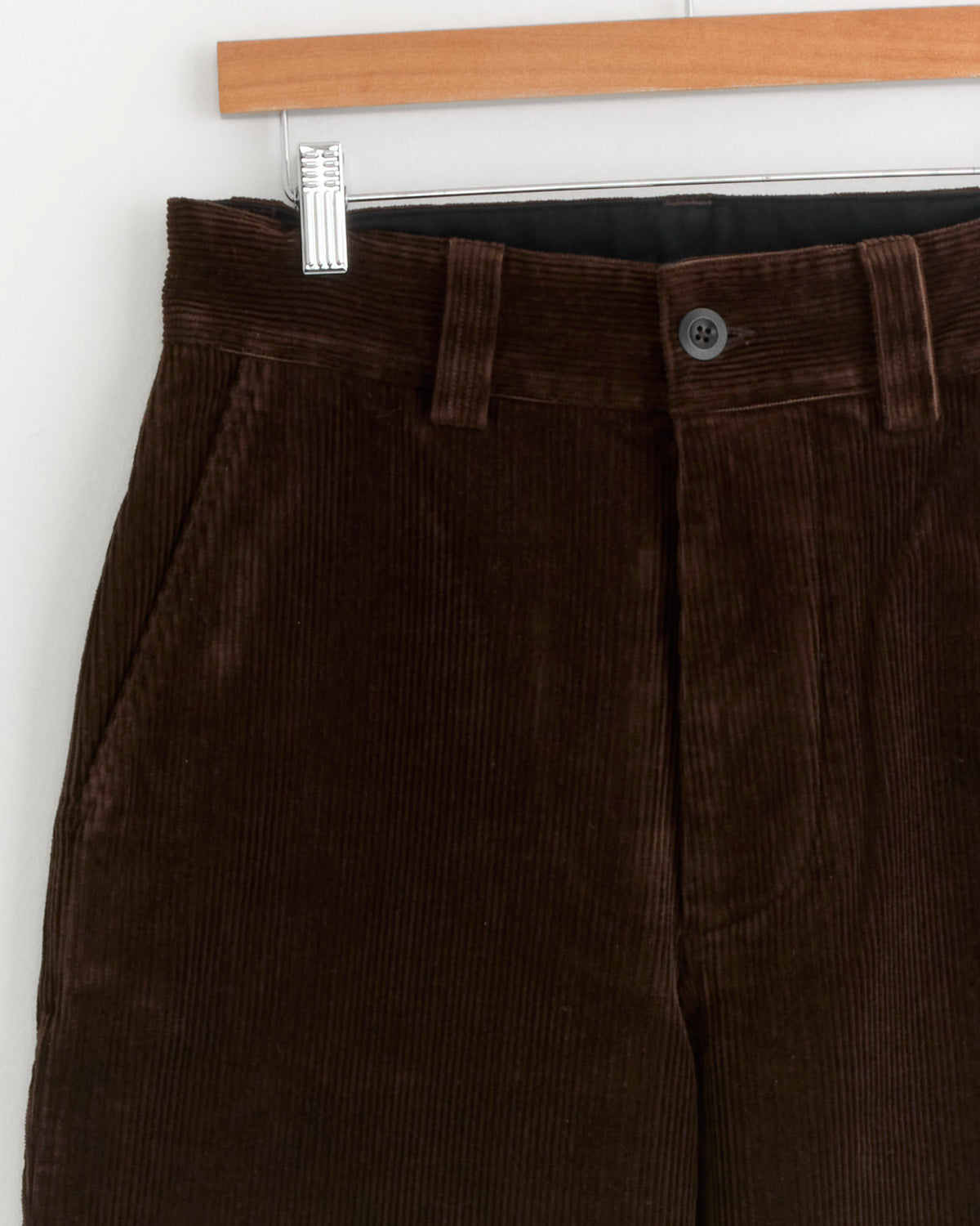 Close-up of the upper part of the Simple Corduroy Trouser in Chestnut by MHL by Margaret Howell, hanging on a wooden hanger against a white wall. Crafted in Portugal from heavyweight cotton, these trousers feature side pockets, belt loops, and a single black button at the waistband.