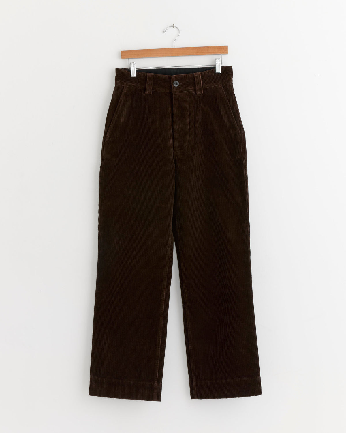 The Simple Corduroy Trouser in Chestnut by MHL by Margaret Howell, crafted from heavyweight cotton with a straight-leg cut, front button and zipper closure, is displayed hanging on a wooden hanger against a plain white background.