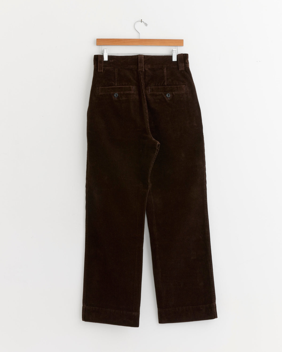 A pair of Simple Corduroy Trousers in Chestnut by MHL by Margaret Howell, made in Portugal from heavyweight cotton corduroy, hangs on a wooden hanger against a white background, displayed from the back.