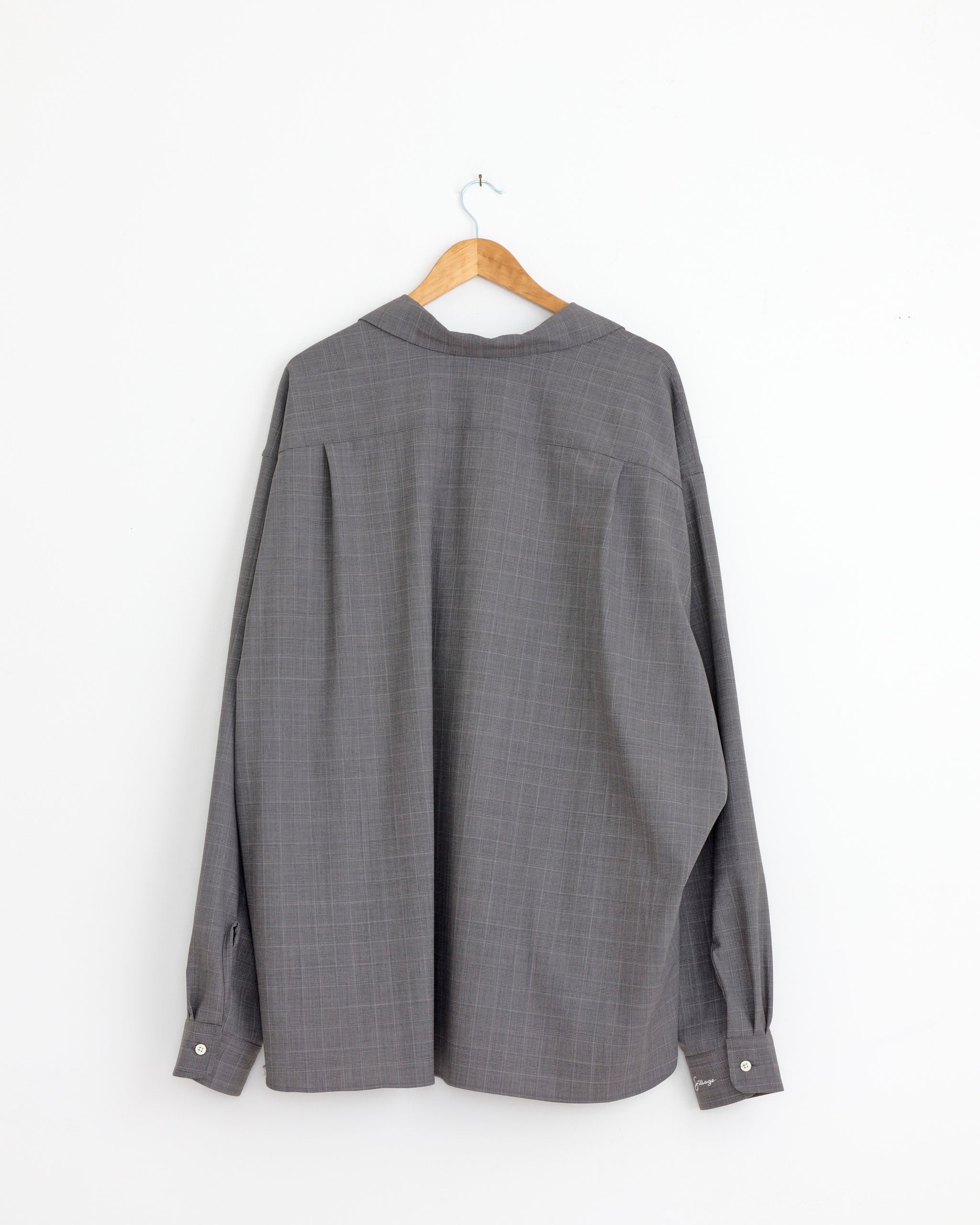 The Sillage Re-Engineered Overshirt in Glen Check, a gray tropical wool long-sleeve shirt, is displayed from the back on a wooden hanger against a plain white wall.