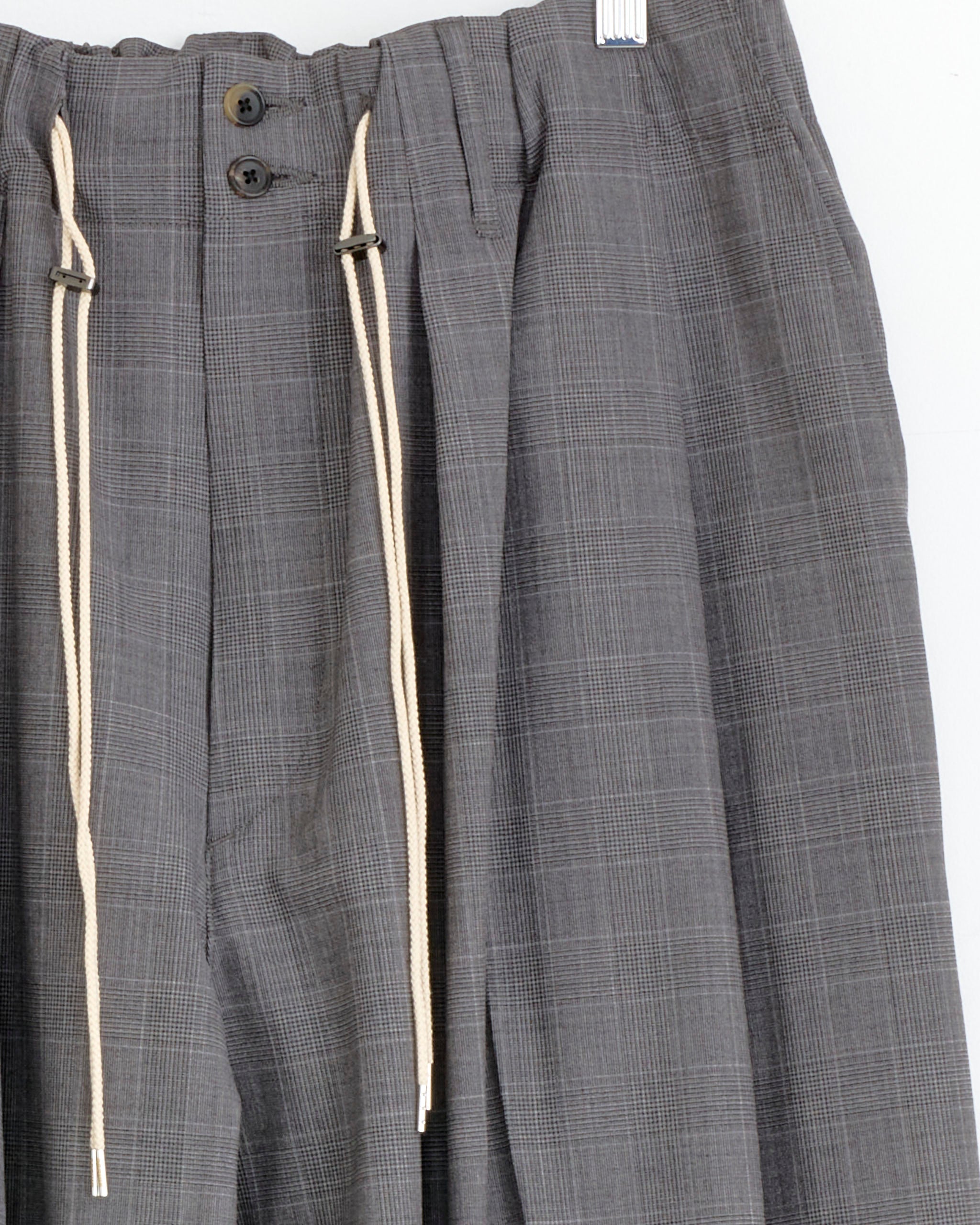 The Sillage Circular Pant in Glen Check features a gray plaid design, high waist, pleats, wide silhouette, two dark buttons, a metal waistband clip, and cream drawstrings down the front.