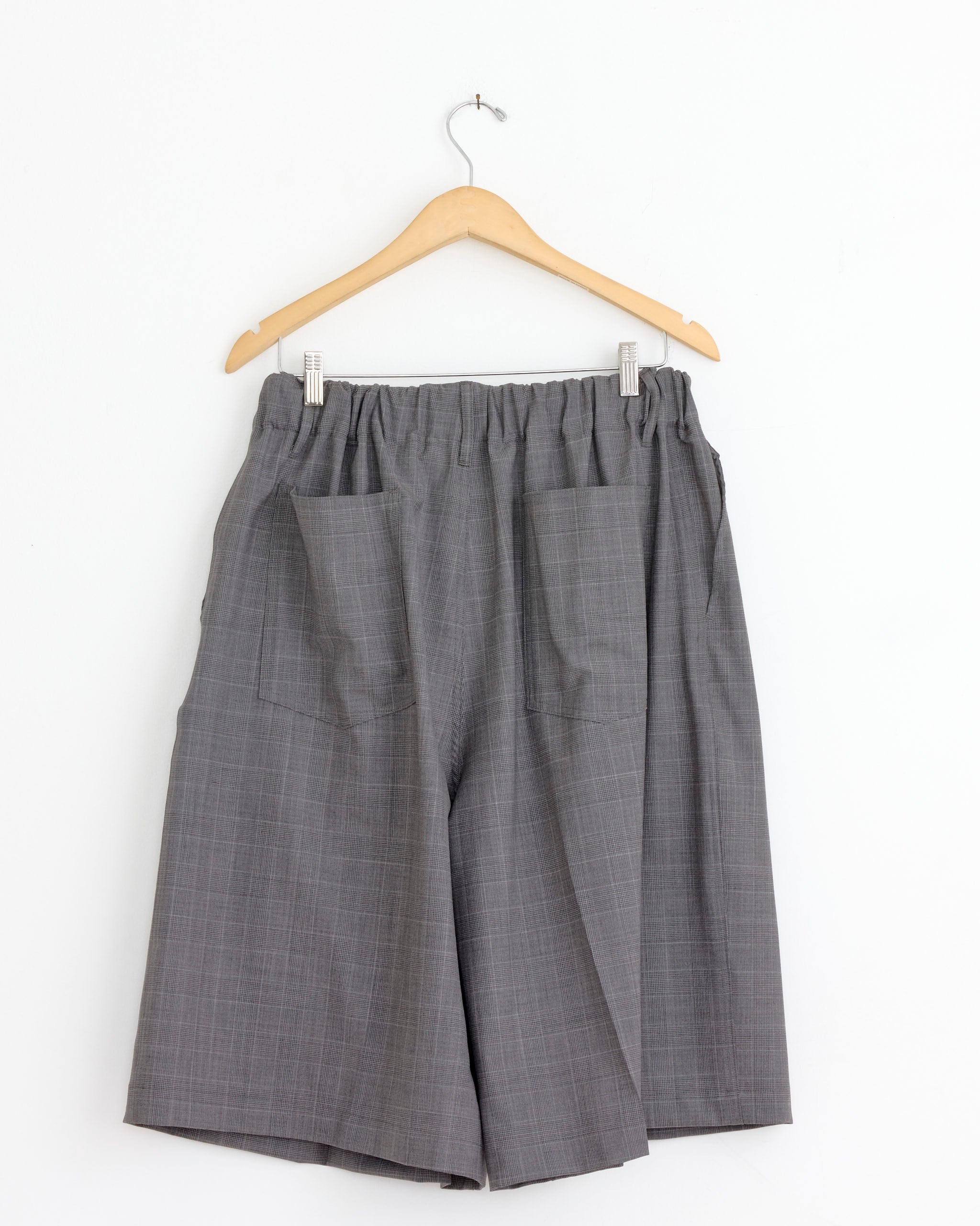The Sillage Circular Short Pant in Glen Check is a loose-fitting gray pant made from tropical wool, featuring an elastic waistband and front patch pockets, shown on a wooden hanger against a white background.