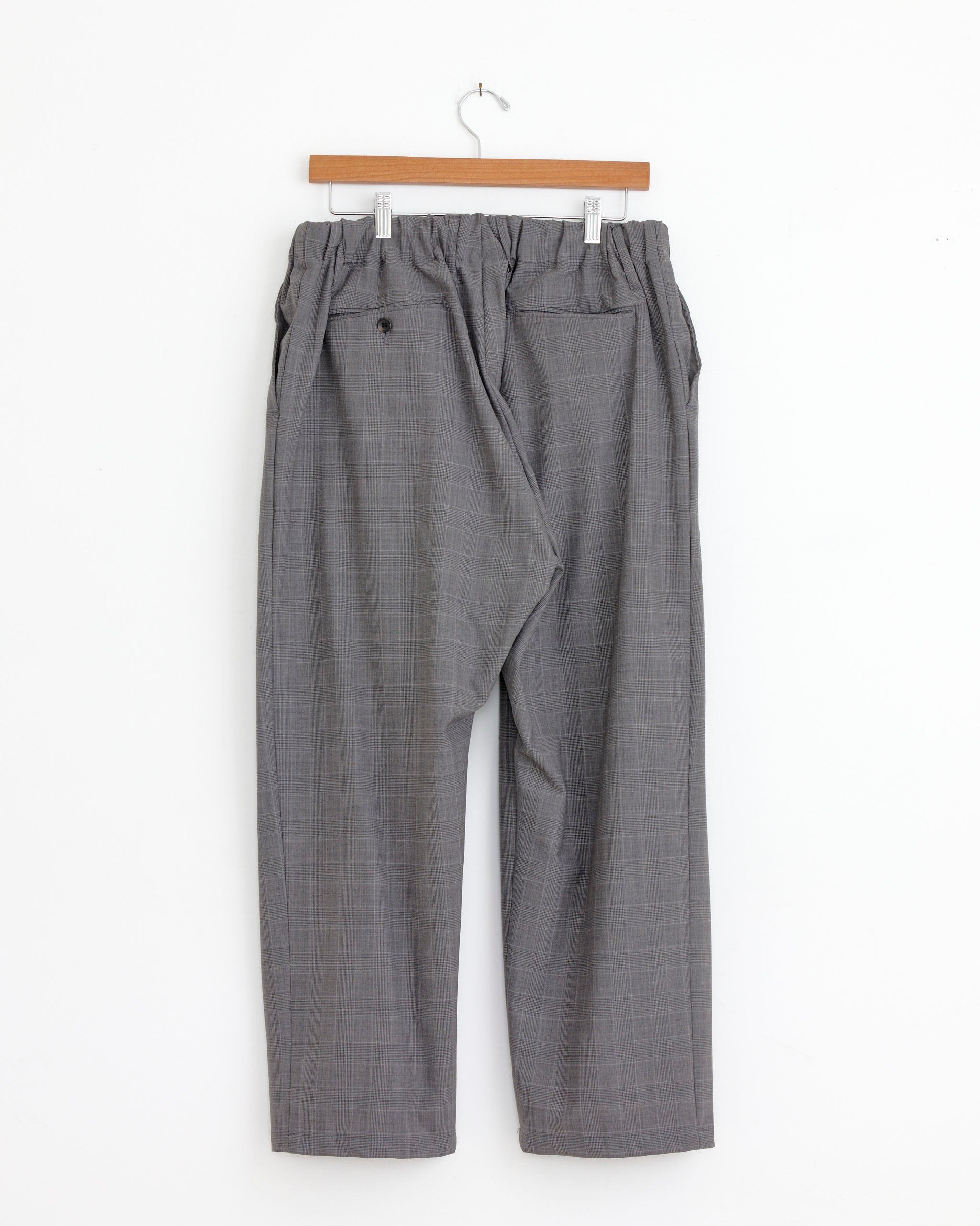 The Sillage Baggy Trouser in Glen Check features a gray subtle-pattern, elastic waistband, and two back pockets, shown on a wooden hanger against a white background. Made in Japan for quality and style.