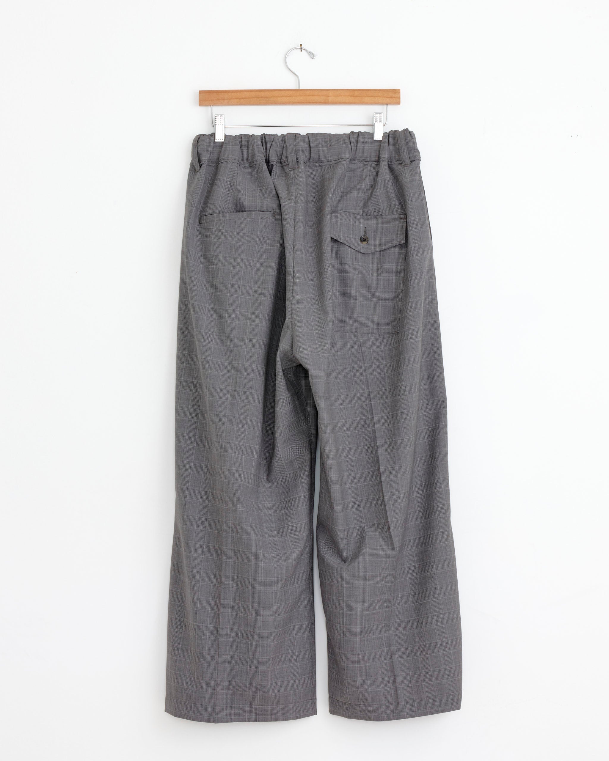 The Sillage Hakama Pant in Glen Check hangs on a wooden hanger against a white wall, shown from the back with an elastic waistband, two back pockets, and a wide straight silhouette for relaxed comfort.