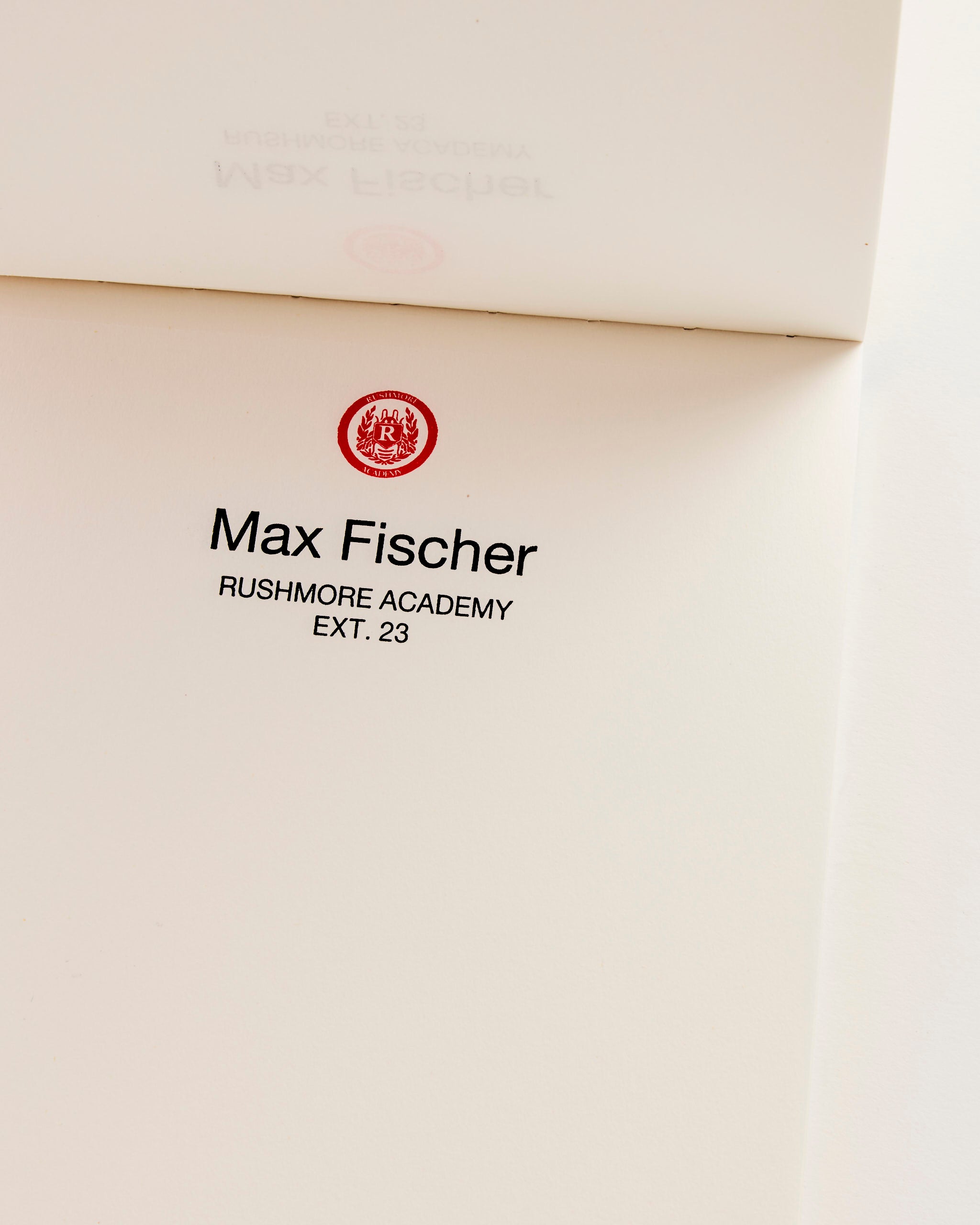The Astier de Villatte Wes Anderson Letter Paper Notepad features an open page with "Max Fischer, Rushmore Academy, EXT. 23" beneath a red emblem on a plain white background, evoking the iconic style of Wes Anderson.
