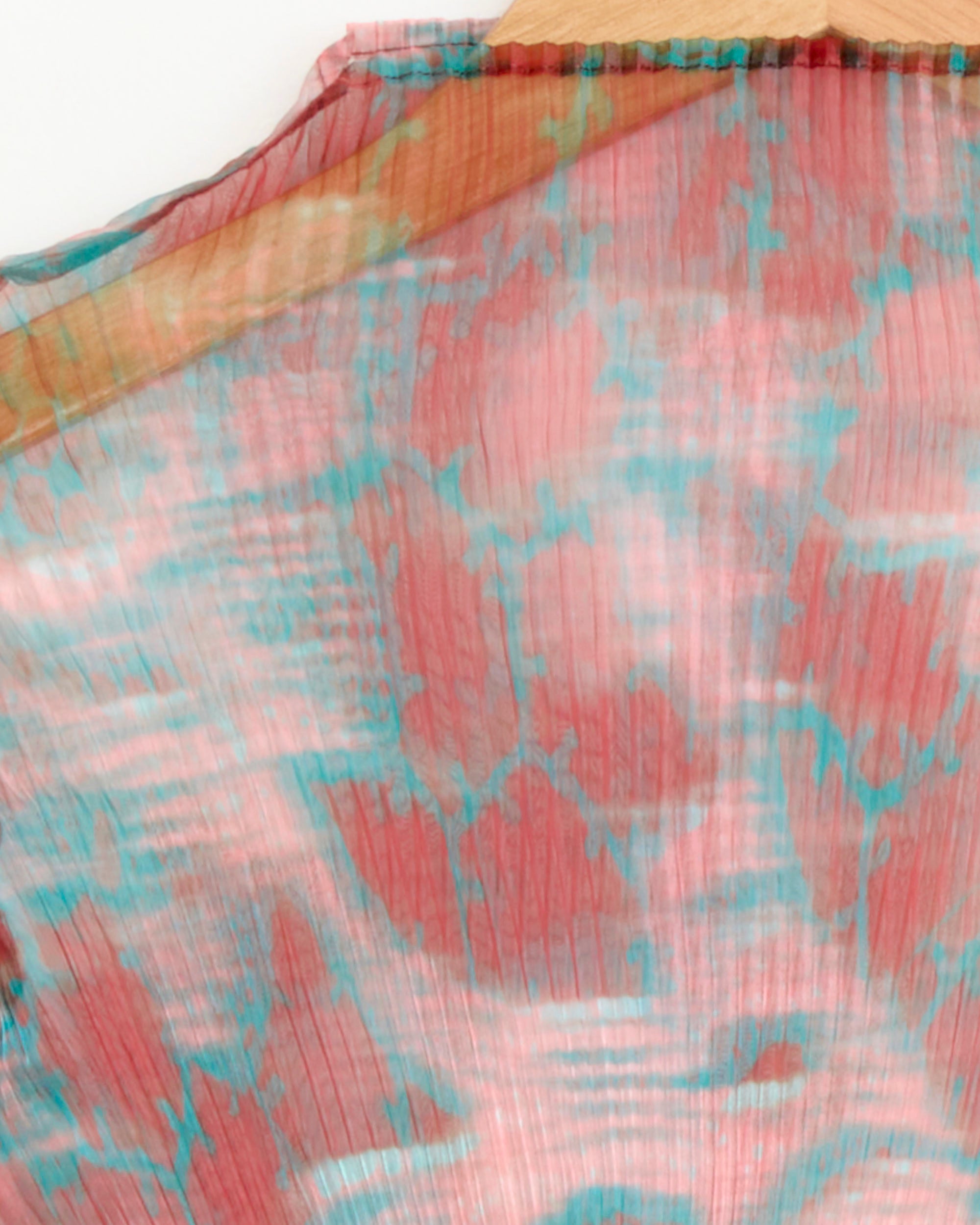 Close-up of Julia Heuer's Lee Top in Gallia, featuring sheer pleated crepe with a watercolor-inspired pattern in coral pink, teal, and white, draped on a wooden hanger.