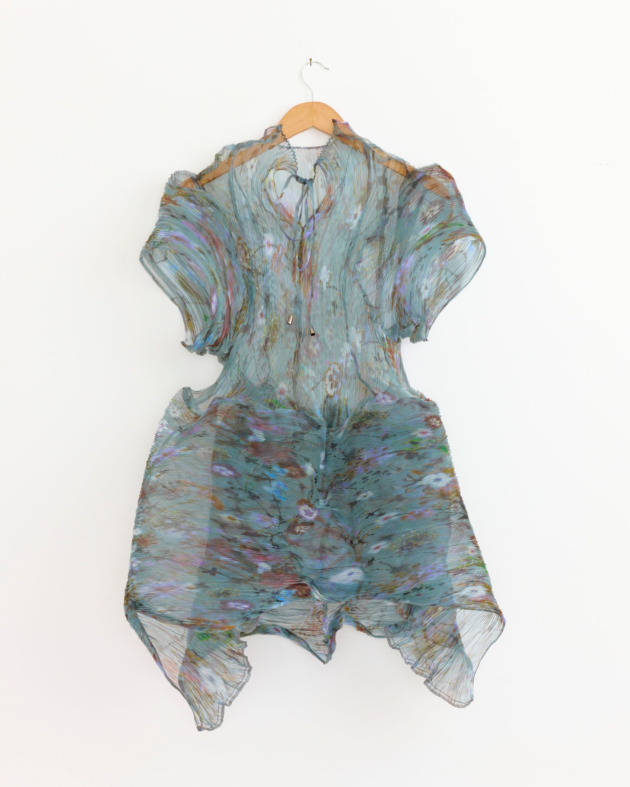 The Thekla Transparent Dress in Frances by Julia Heuer, featuring a wavy, multicolored pastel floral pattern, is a sheer, short-sleeved knee-length design displayed on a wooden hanger against a white background.