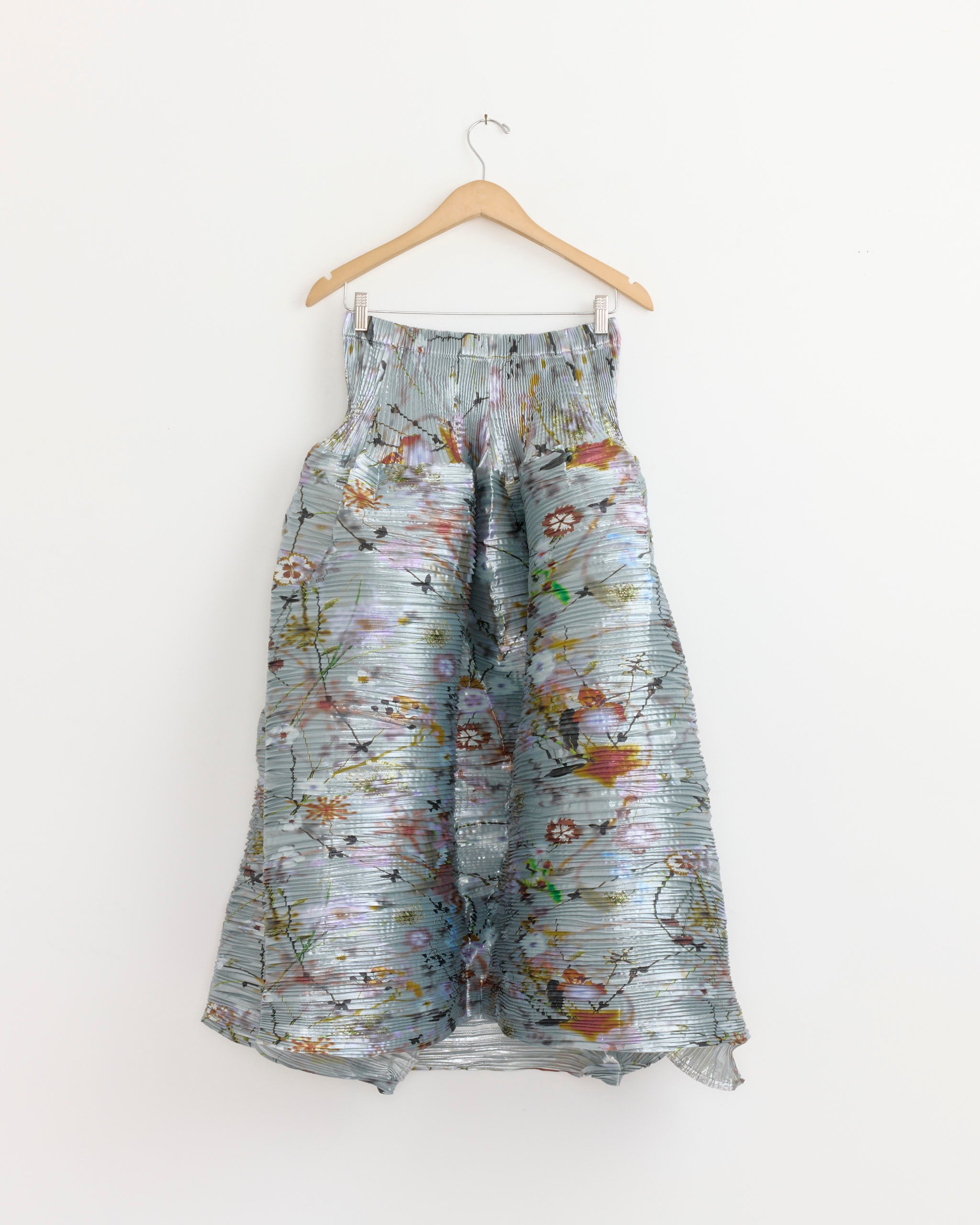 The Julia Heuer Kenza Skirt in Frances, a high-waisted, long skirt with a voluminous silhouette and hand-pleated arashi shibori fabric adorned with a colorful floral print, hangs on a wooden hanger against a white background.