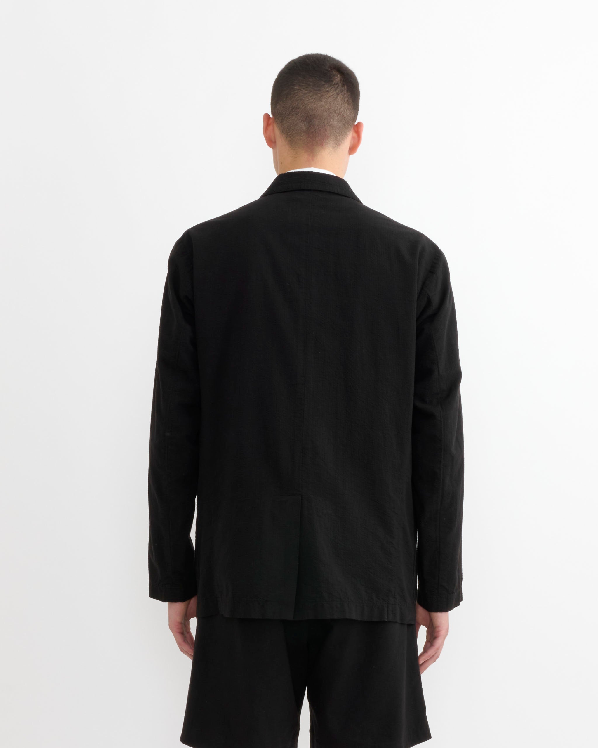 A person with short hair stands against a plain white background, facing away from the camera, wearing SMOCK's Blazer in Japanese Seersucker Black and black shorts.