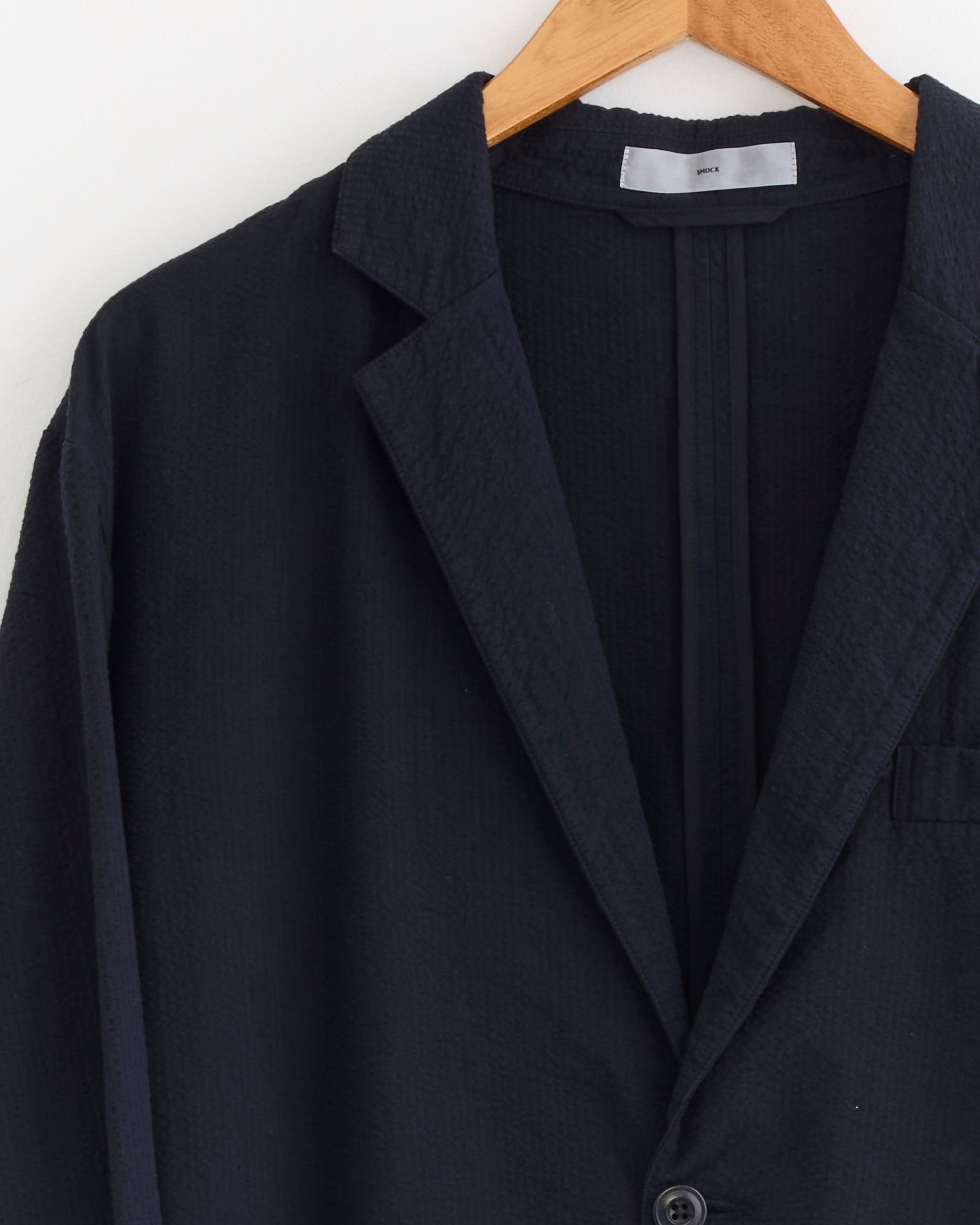 A close-up of the SMOCK Blazer in Japanese Seersucker Navy hangs on a wooden hanger against a white background, showing only the upper half and left sleeve of this unconstructed blazer.