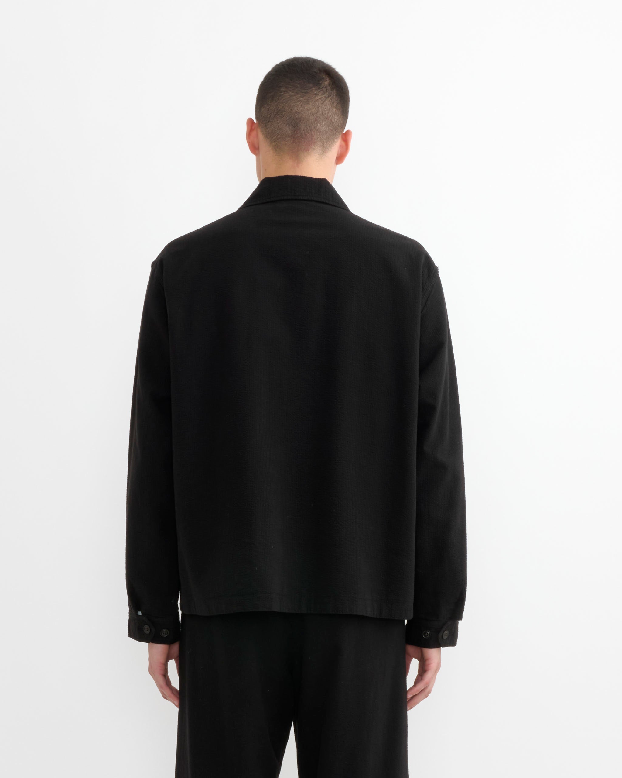 A person with short hair faces away from the camera, wearing SMOCK's Knoll Jacket in Japanese Seersucker Black and black pants, set against a plain white background.