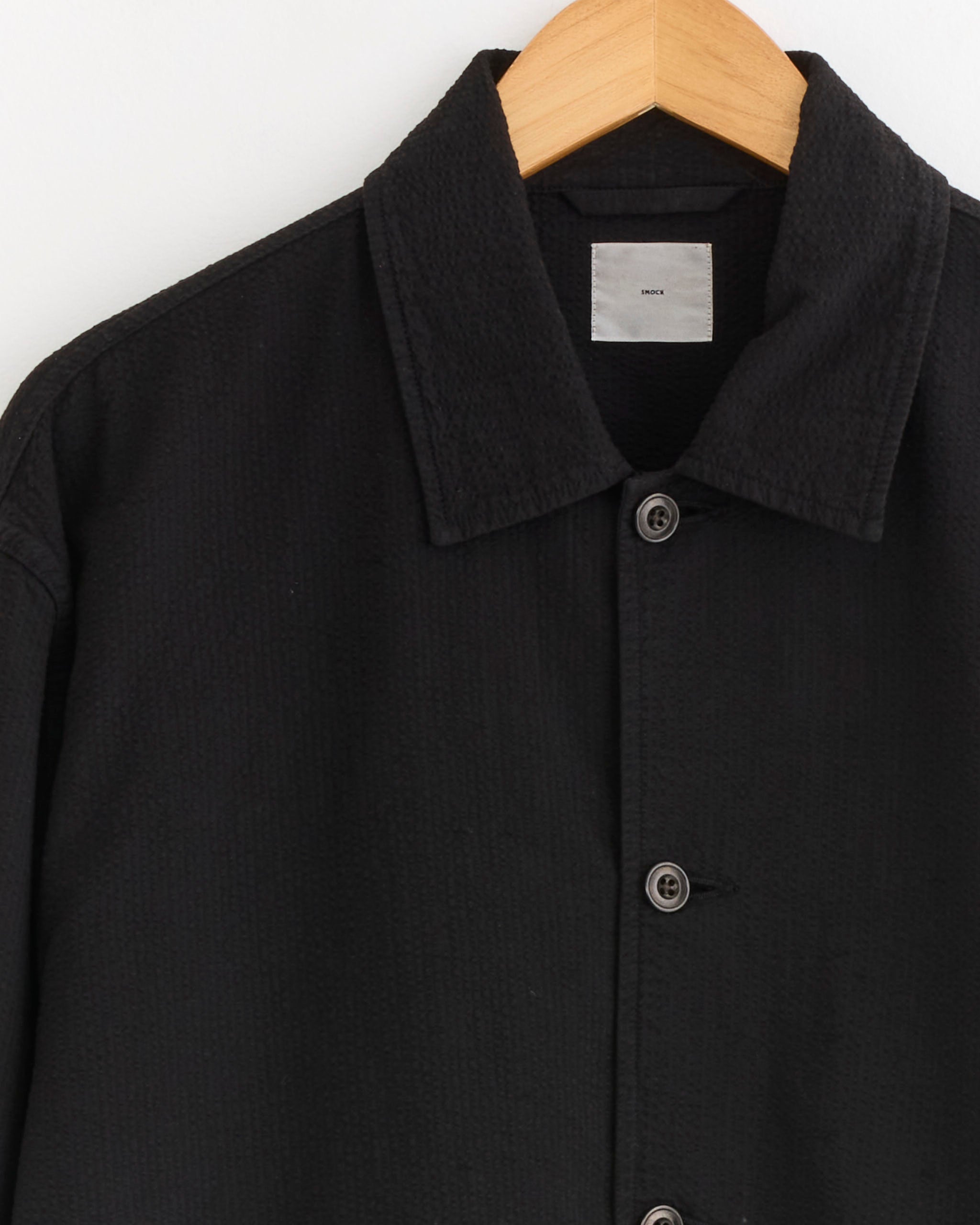A close-up of the SMOCK Knoll Jacket in Japanese Seersucker Black, displayed on a wooden hanger, highlights its collar, three buttons, and a partial white label—a modern update to the classic coaches jacket.