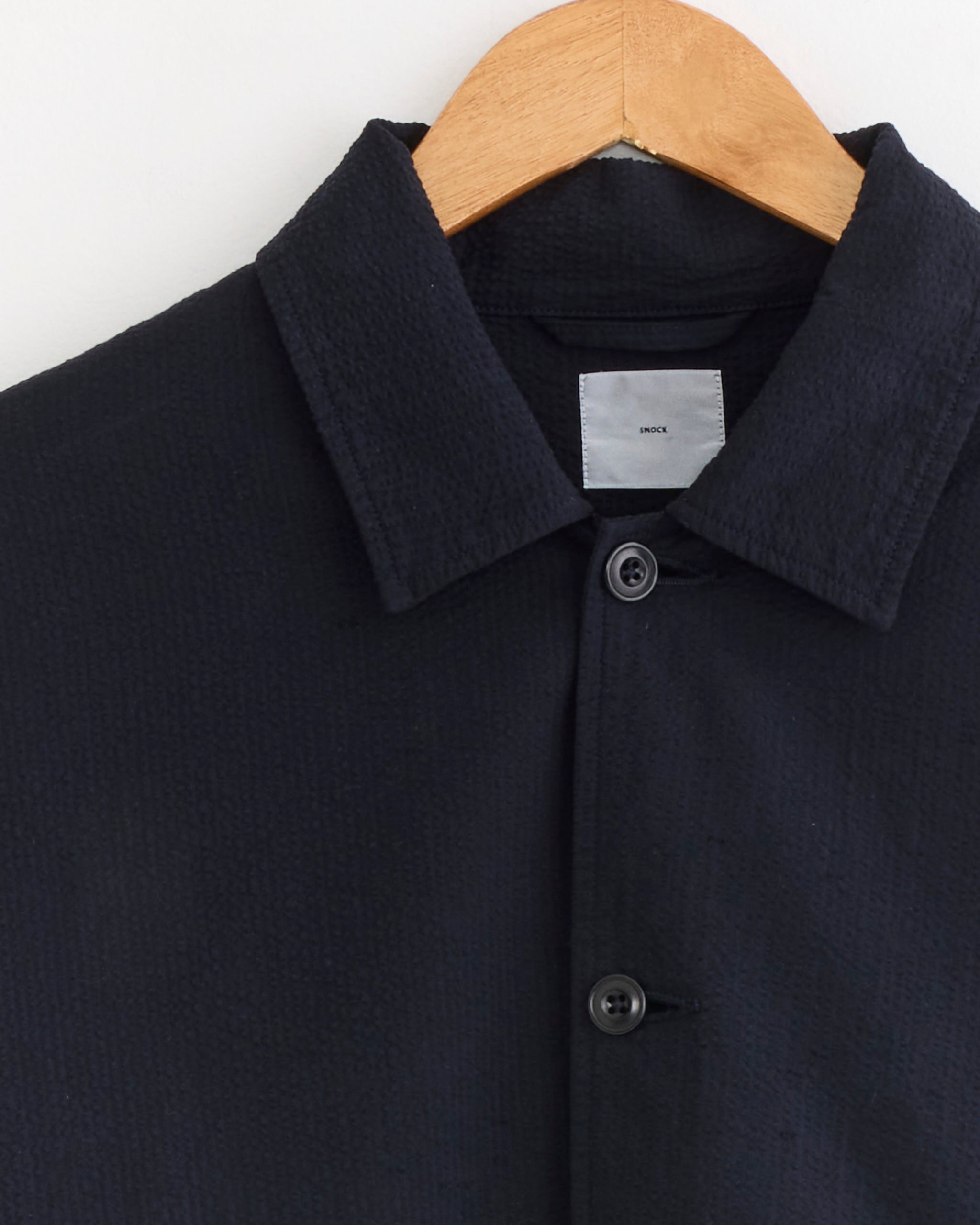 A SMOCK Knoll Jacket in Japanese Seersucker Navy hangs on a wooden hanger against a white background, with the top button fastened and a small white label visible inside the collar.