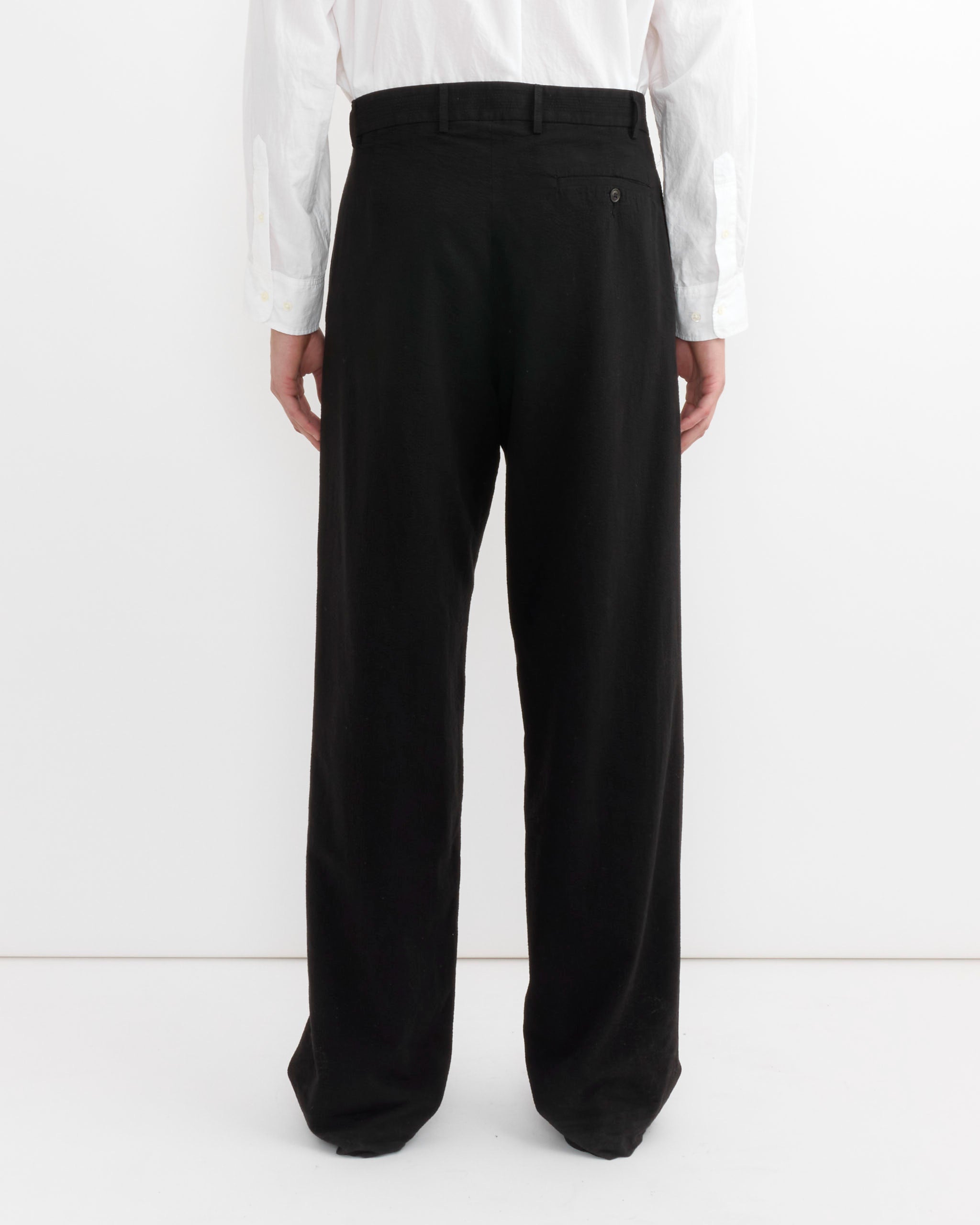 A person stands facing away from the camera, wearing a white long-sleeve shirt and SMOCK’s Lakeside Trouser in Japanese Seersucker Black with a single back pocket, against a plain white background.