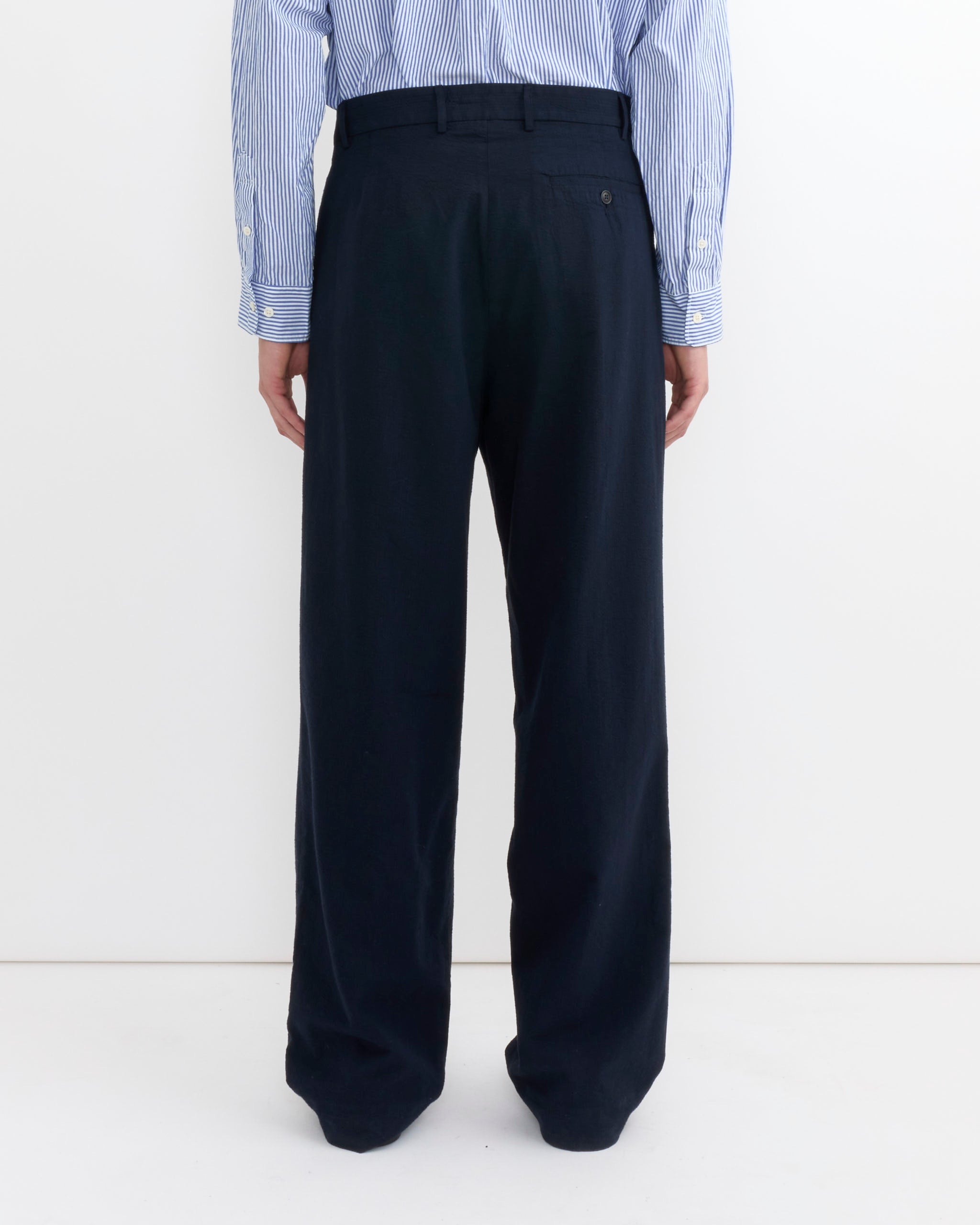 A person stands facing away against a plain white background, wearing a blue and white striped shirt and SMOCK's Lakeside Trouser in Japanese Seersucker Navy.