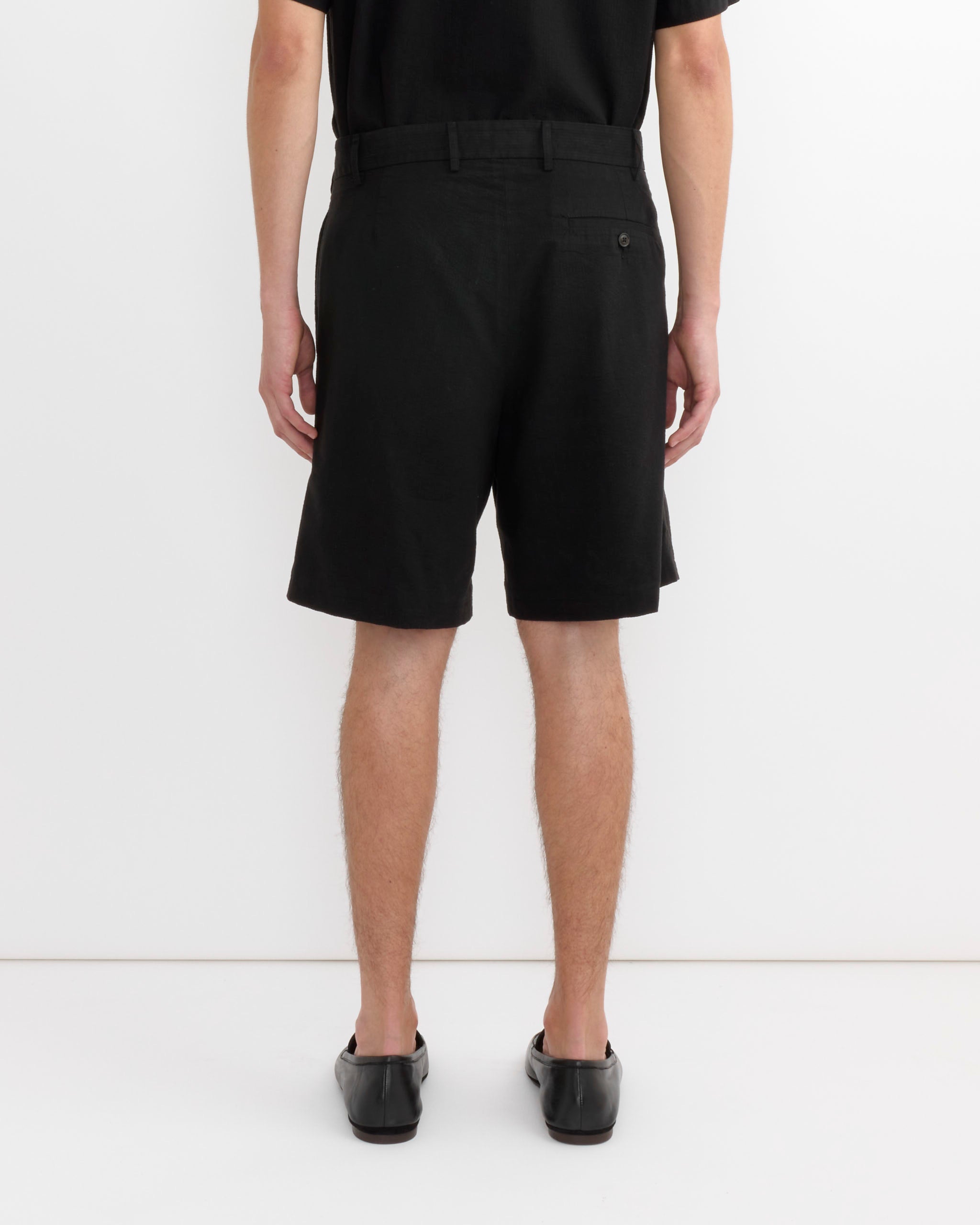 A person stands facing away, arms at their sides, wearing a black shirt, SMOCK Arroyo Short in Japanese Seersucker Black made from lightweight cotton lyocell, and black shoes against a plain white background.