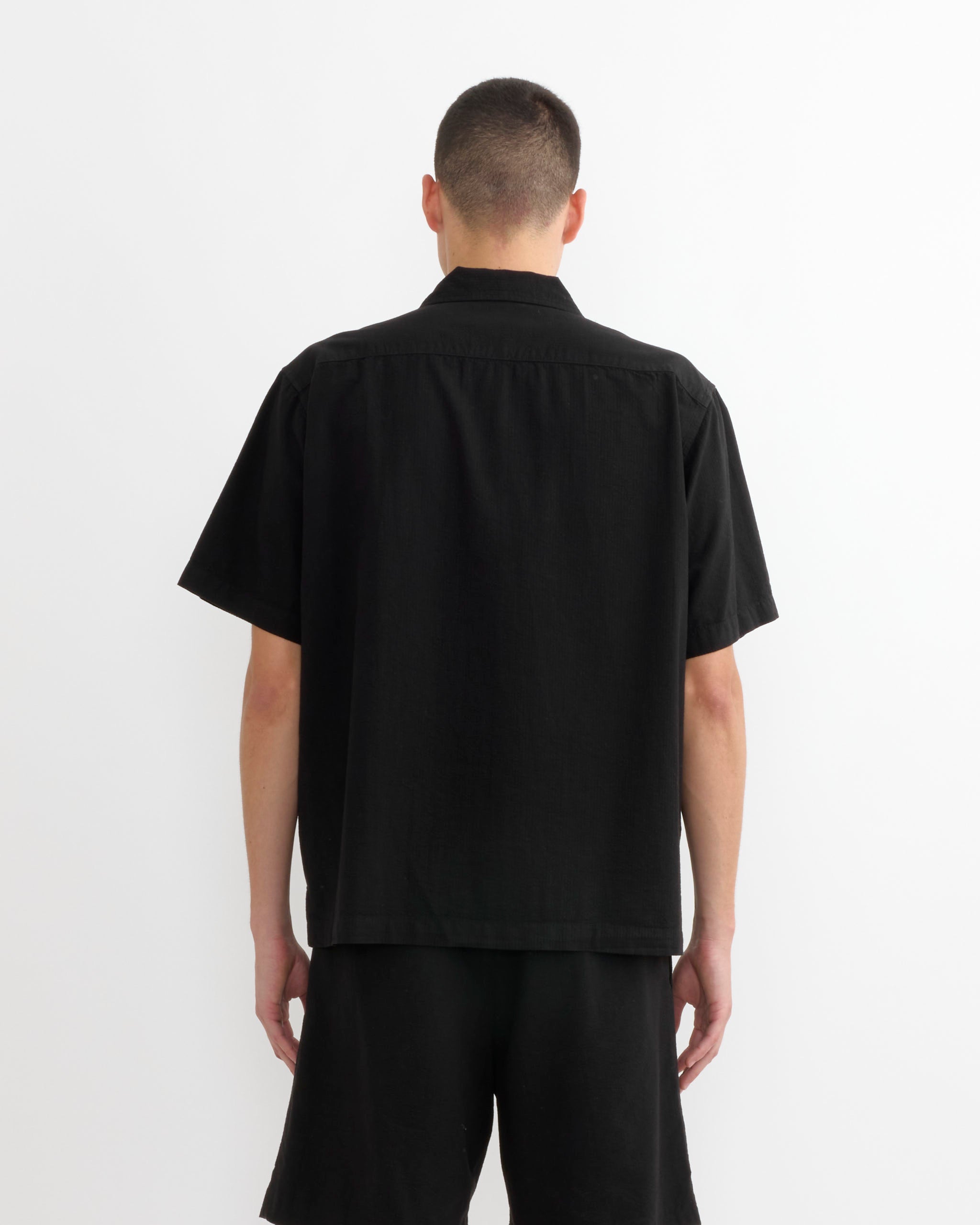 A person with short hair, facing away from the camera, wears the SMOCK Cardiff Shirt in Japanese Seersucker Black and black shorts, set against a plain white background.