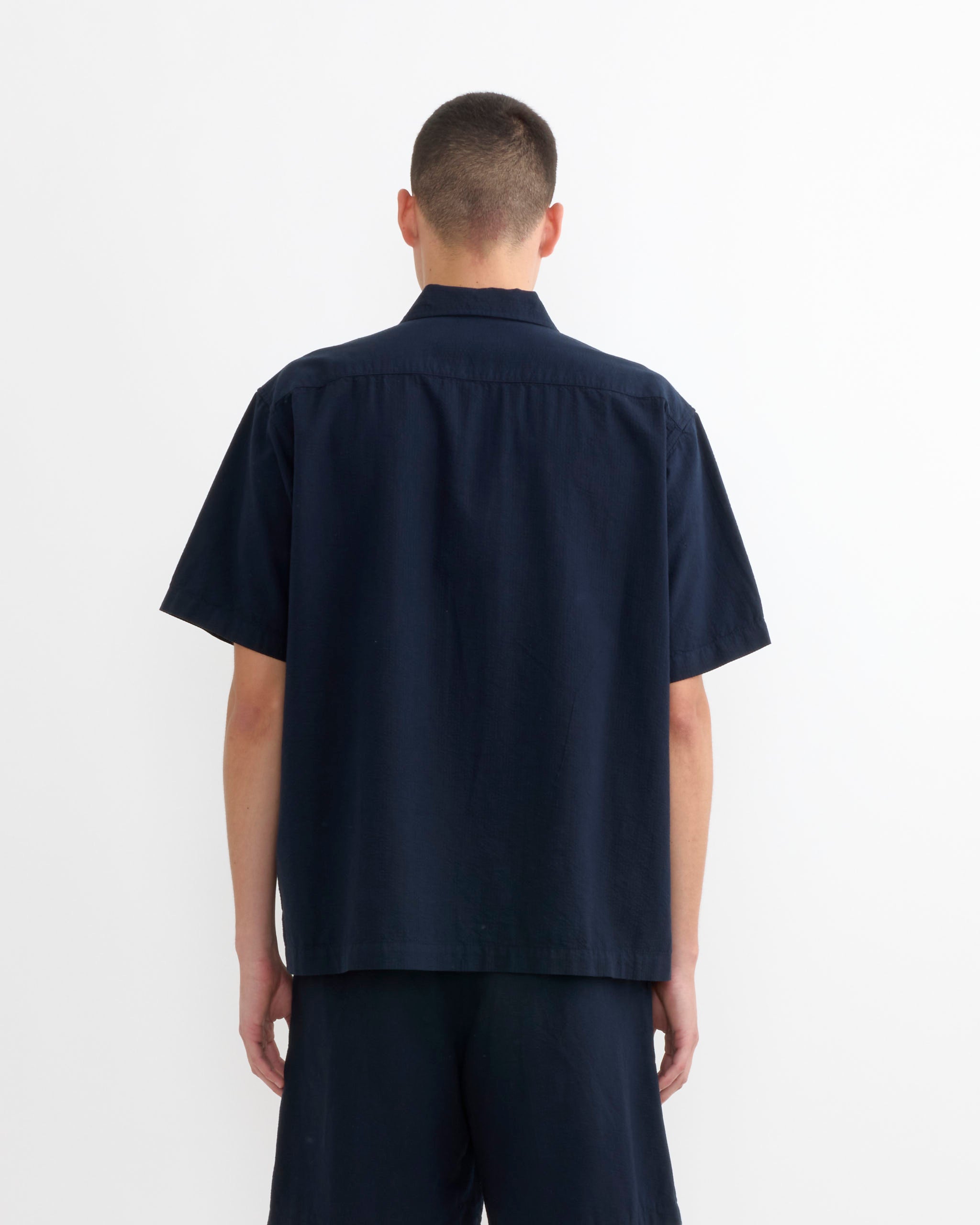 A person with short hair faces away from the camera wearing the SMOCK Cardiff Shirt in Japanese Seersucker Navy, paired with matching shorts, standing against a plain white background.