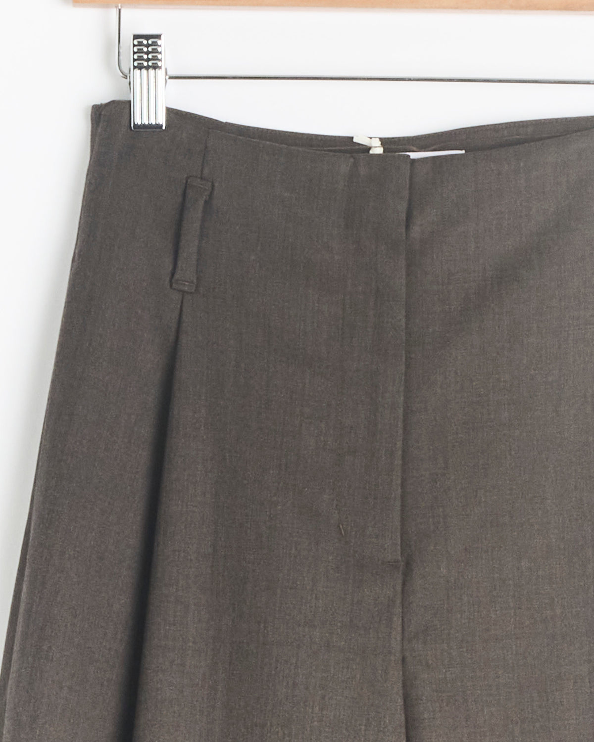 Close-up of the Amomento Straight Tuck Pant in Brown hanging on a wooden hanger, highlighting the waistband, belt loop, zipper, and front pleat with subtle tuck detailing.