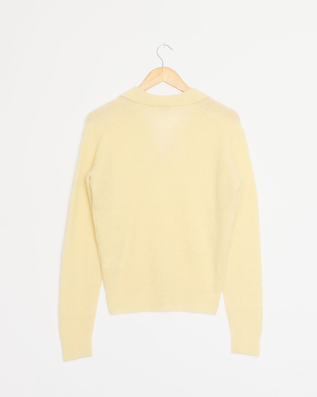 An ecru long-sleeve Baby Suri Alpaca Skipper Polo by Auralee is displayed on a wooden hanger against a plain white background, shown from the back. This luxurious piece is expertly crafted and proudly made in Japan.