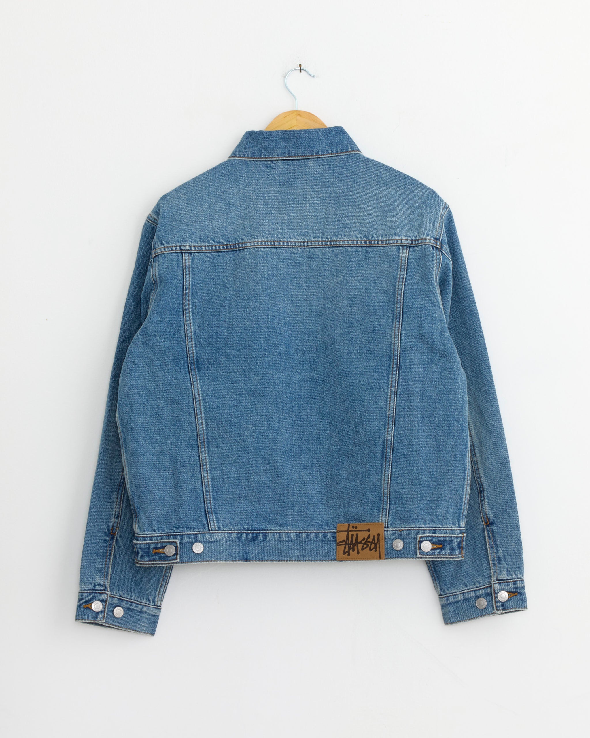 The Stussy Lined Denim Trucker Jacket in Indigo Stone Wash hangs on a wooden hanger against a white wall, shown from the back with a brown Stussy label above the waistband.