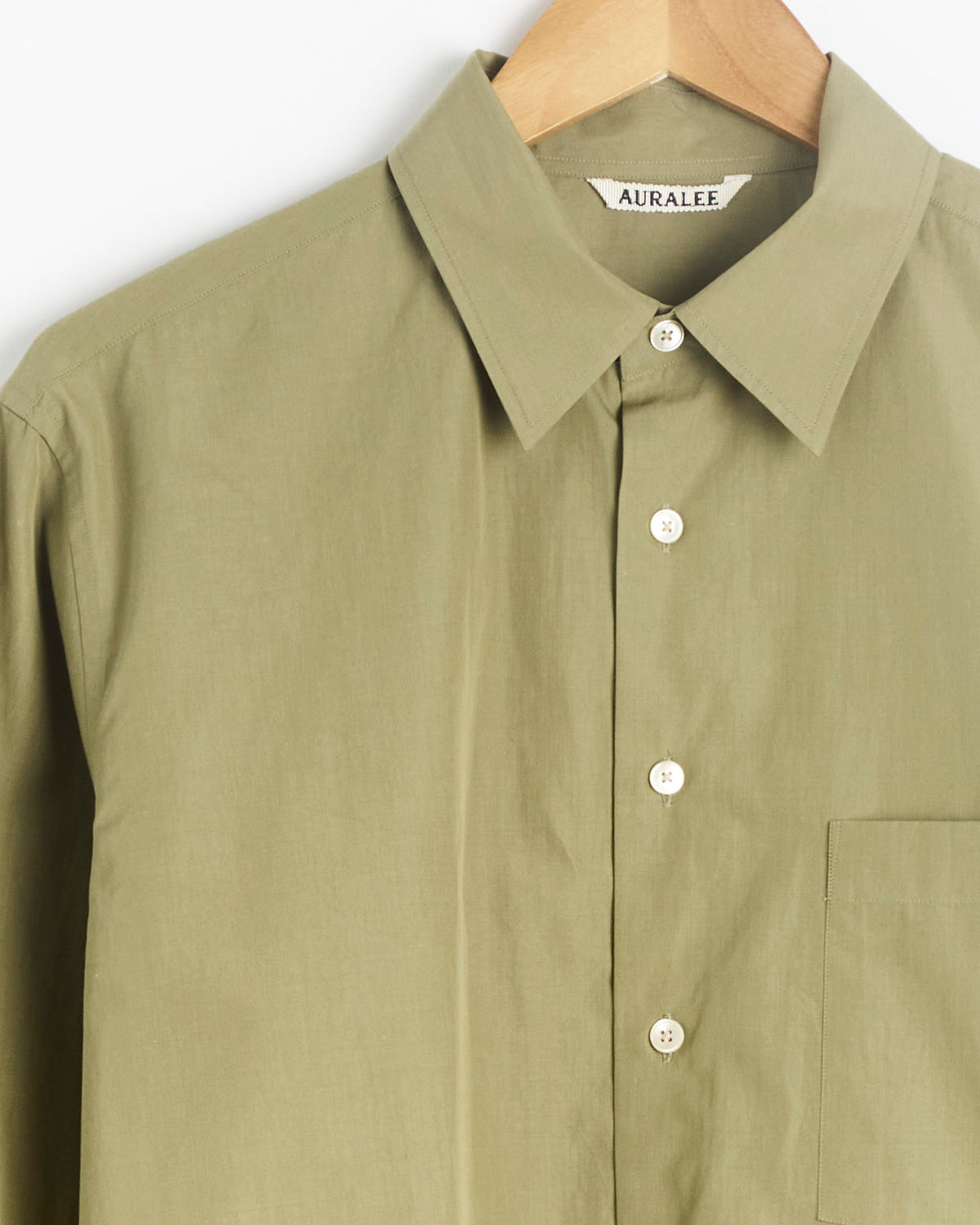 Finx Light Weather Chambray Shirt in Olive Brown by Auralee, shown on a wooden hanger with a pointed collar, white buttons, a visible chest pocket, and an Auralee label inside the collar—crafted with expert precision in Japan.
