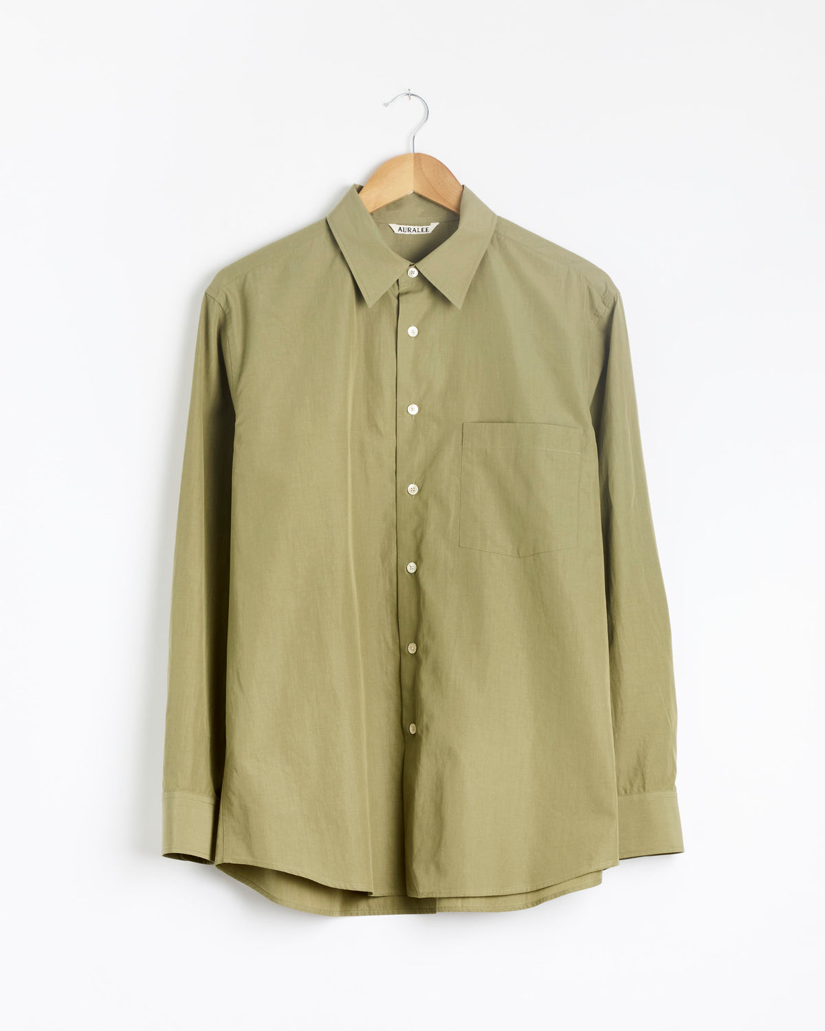 An Auralee Finx Light Weather Chambray Shirt in olive brown, featuring long sleeves, a collar, and a chest pocket, is displayed on a wooden hanger against a white background.