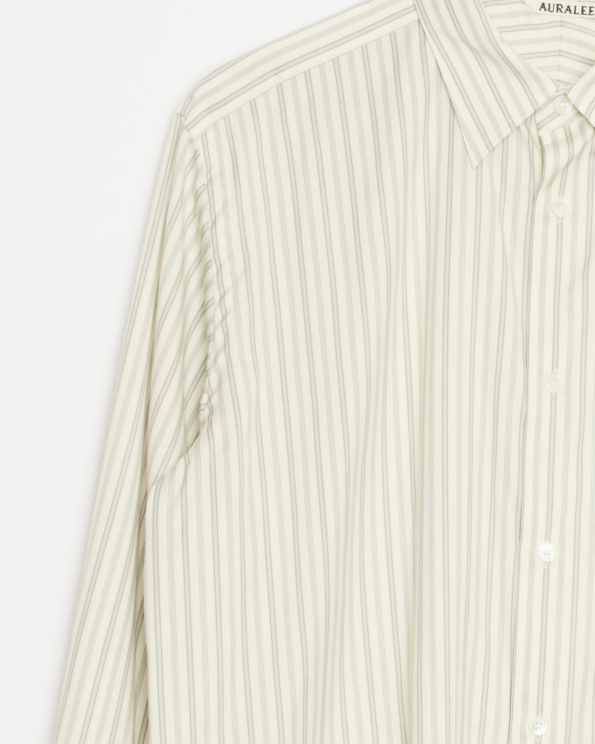 A close-up of Auralee’s Cotton Silk Stripe Shirt in Light Grey showcases thin vertical stripes in beige and white. Expertly crafted from luxurious Egyptian Giza cotton, this shirt features a collar with the brand name Auralee visible on the tag. Made in Japan.