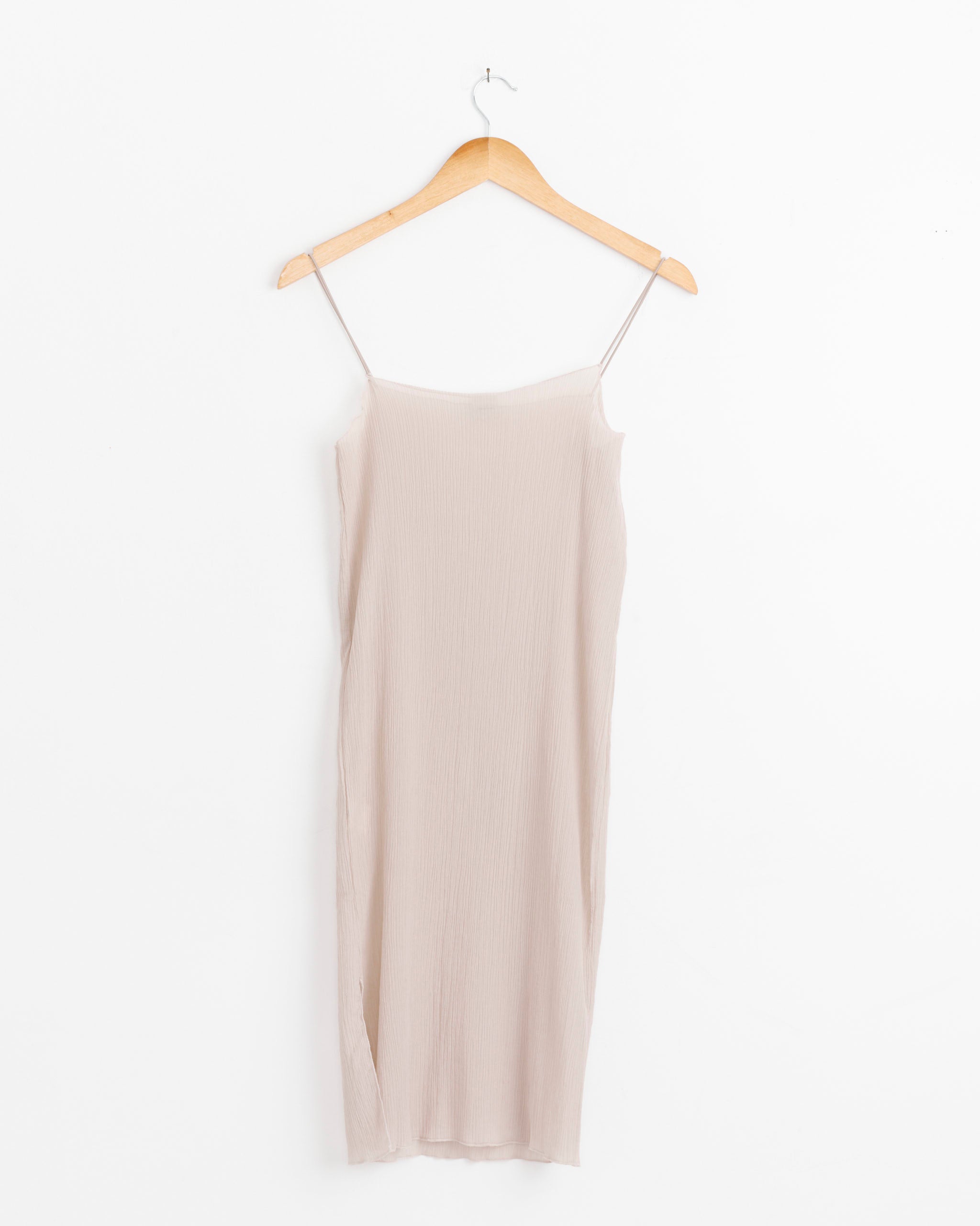 The Sti Slip Dress in Sylva Beige by Baserange, a light beige sleeveless organic cotton dress with thin spaghetti straps, hangs on a wooden hanger against a plain white background.